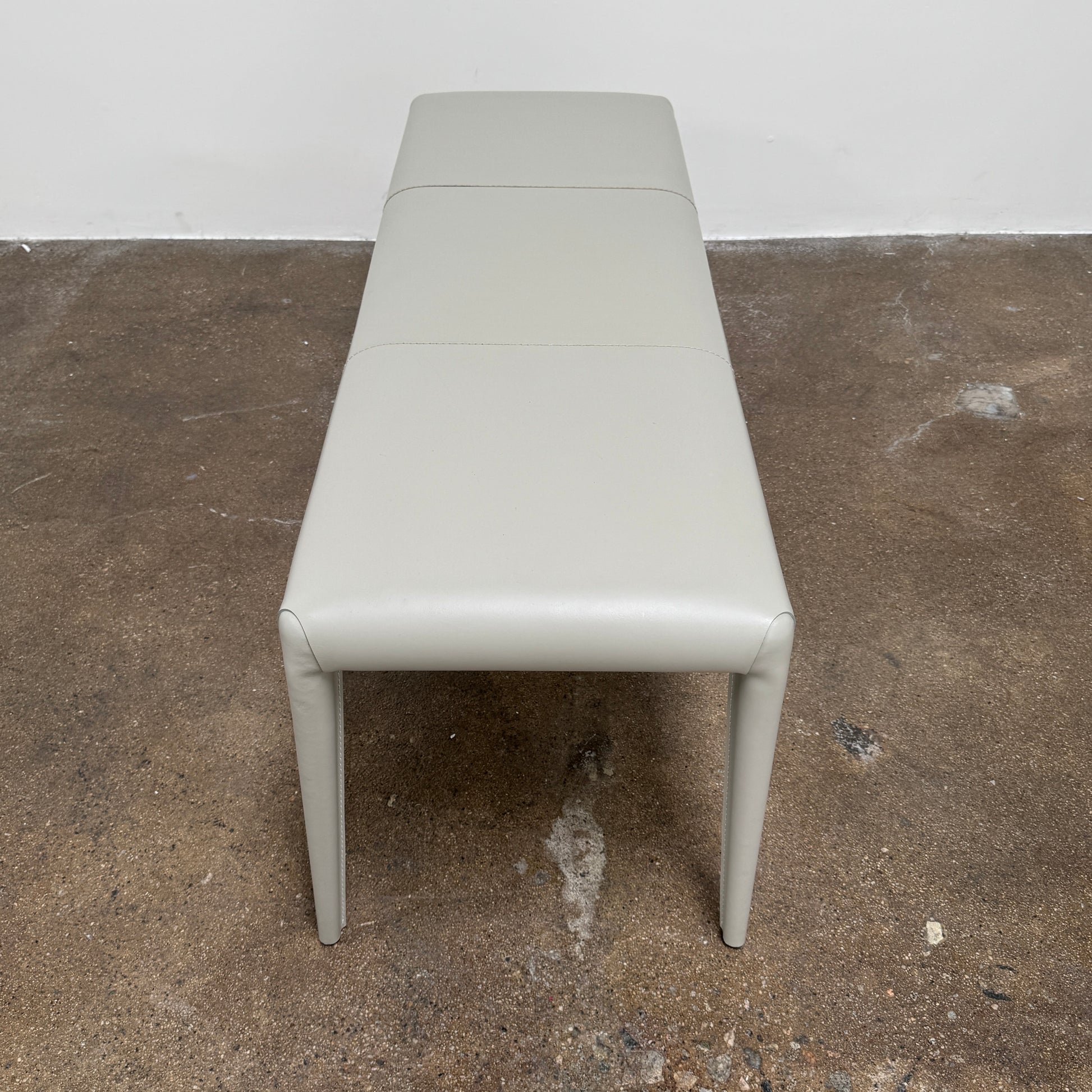 A minimalist, light gray Frag Vella bench by Frag, featuring a padded seat and four legs and handmade in Italy, is positioned against a plain white wall on a brown concrete floor.