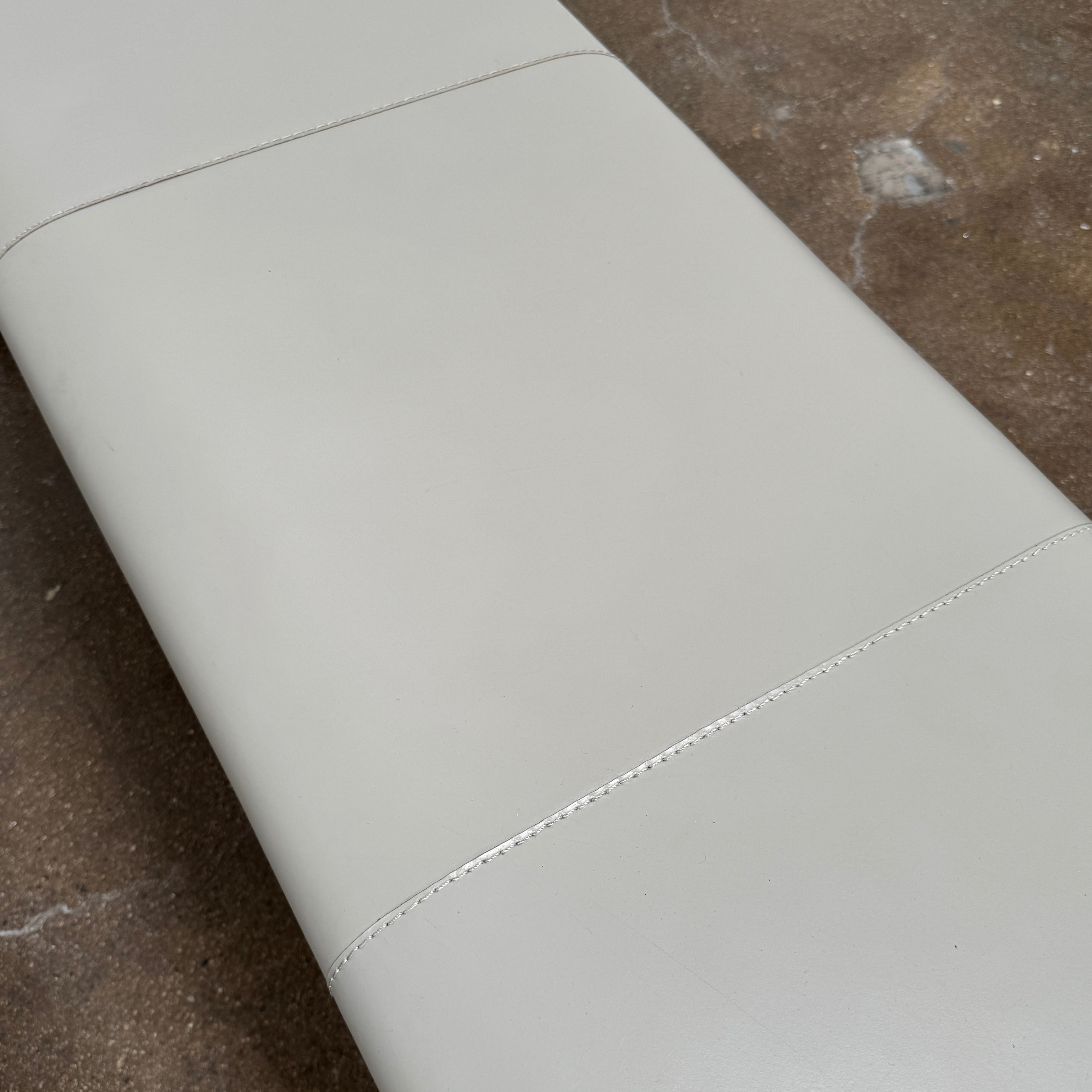 A minimalist, light gray Frag Vella bench by Frag, featuring a padded seat and four legs and handmade in Italy, is positioned against a plain white wall on a brown concrete floor.