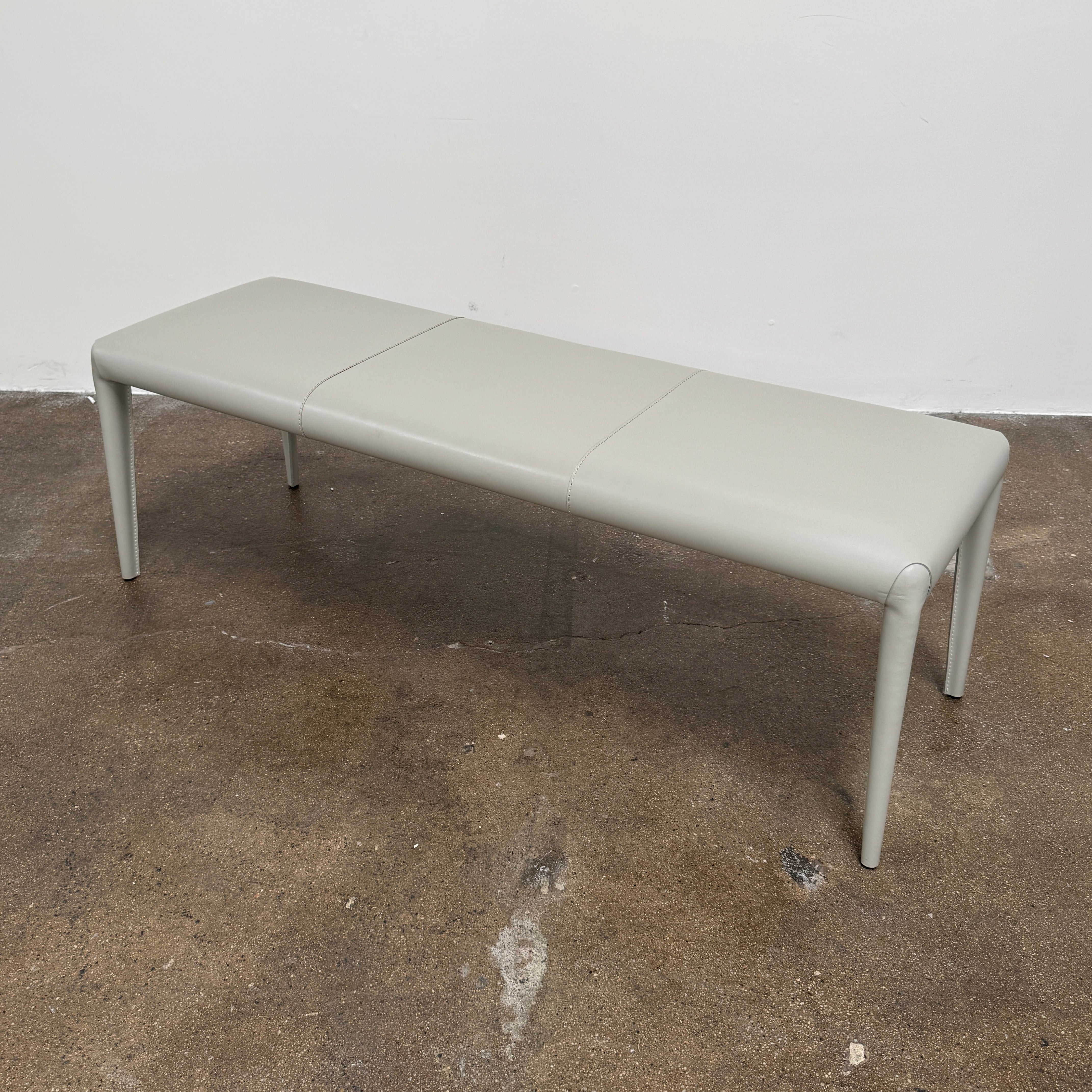 A minimalist, light gray Frag Vella bench by Frag, featuring a padded seat and four legs and handmade in Italy, is positioned against a plain white wall on a brown concrete floor.