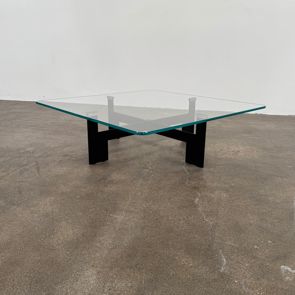 The Minotti Ellis Coffee Table Clear Glass by Minotti features a square clear glass top and a sculptural black metal base, elegantly positioned on a brown concrete floor against a plain white wall.