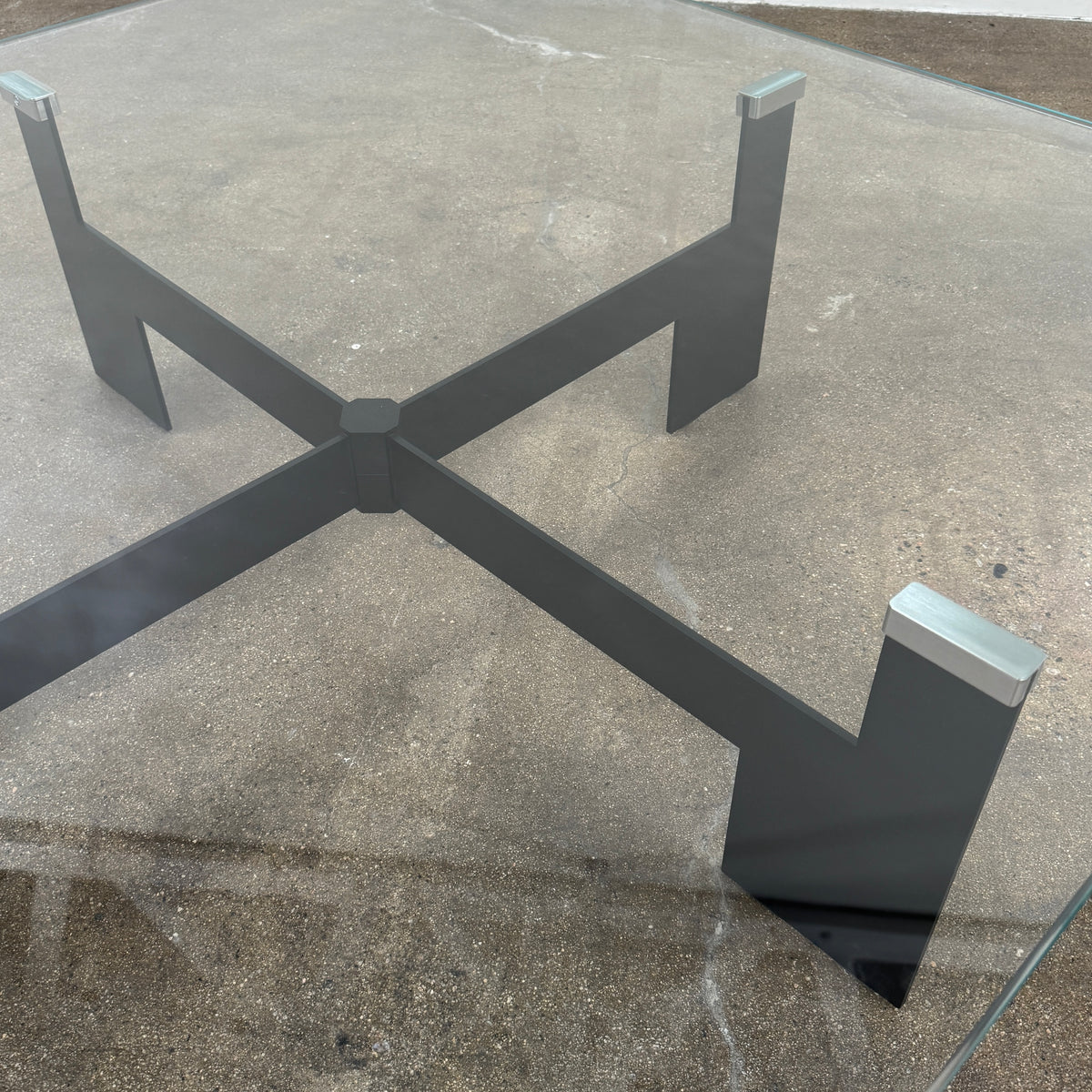 The Minotti Ellis Coffee Table Clear Glass by Minotti features a square clear glass top and a sculptural black metal base, elegantly positioned on a brown concrete floor against a plain white wall.