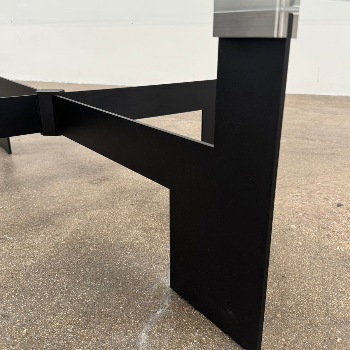 The Minotti Ellis Coffee Table Clear Glass by Minotti features a square clear glass top and a sculptural black metal base, elegantly positioned on a brown concrete floor against a plain white wall.