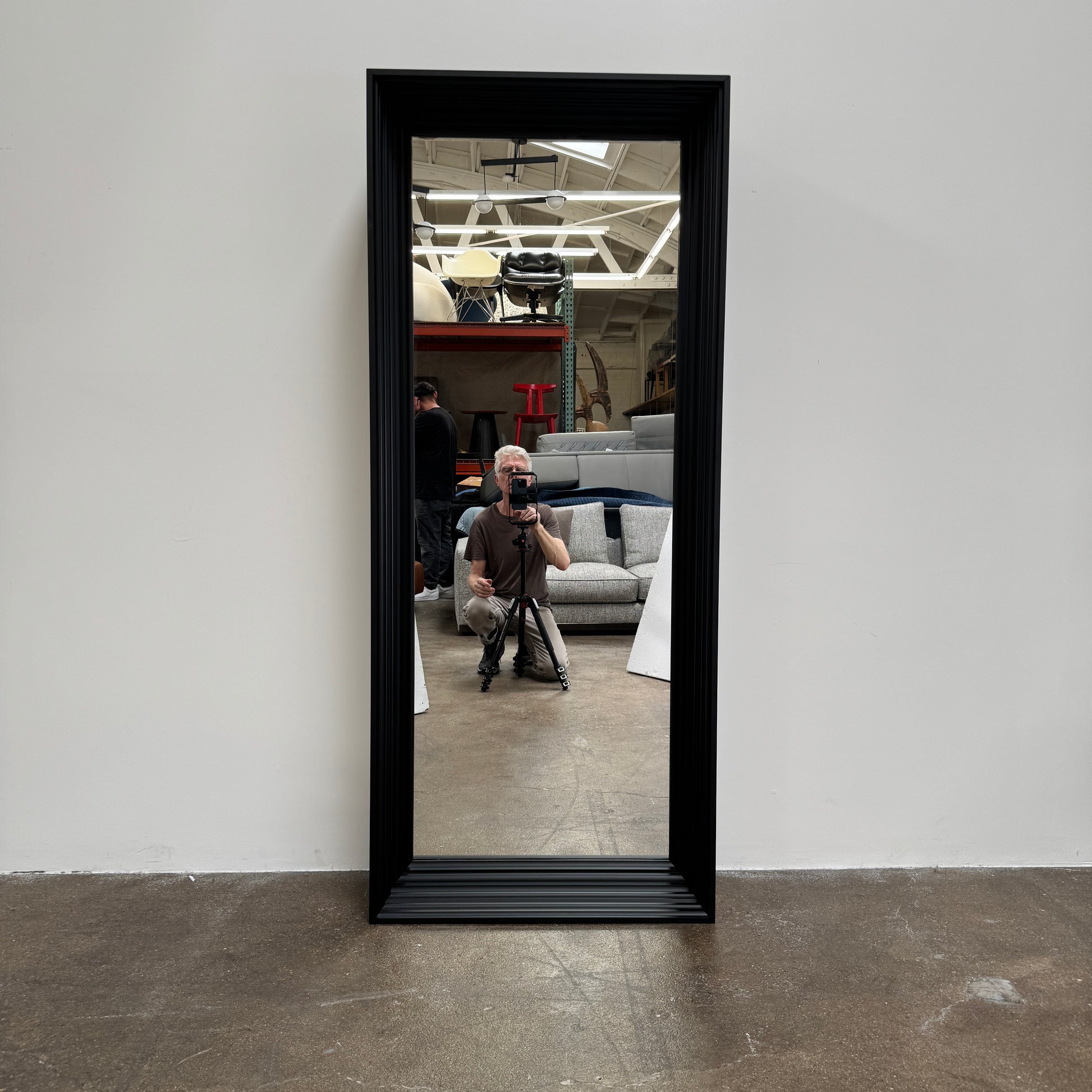 A person kneels in front of the Moooi Frame Mirror by Moooi—a large, rectangular mirror with a sleek black aluminum frame—while taking a photo. The mirror reflects furniture and industrial shelving within a well-lit room.