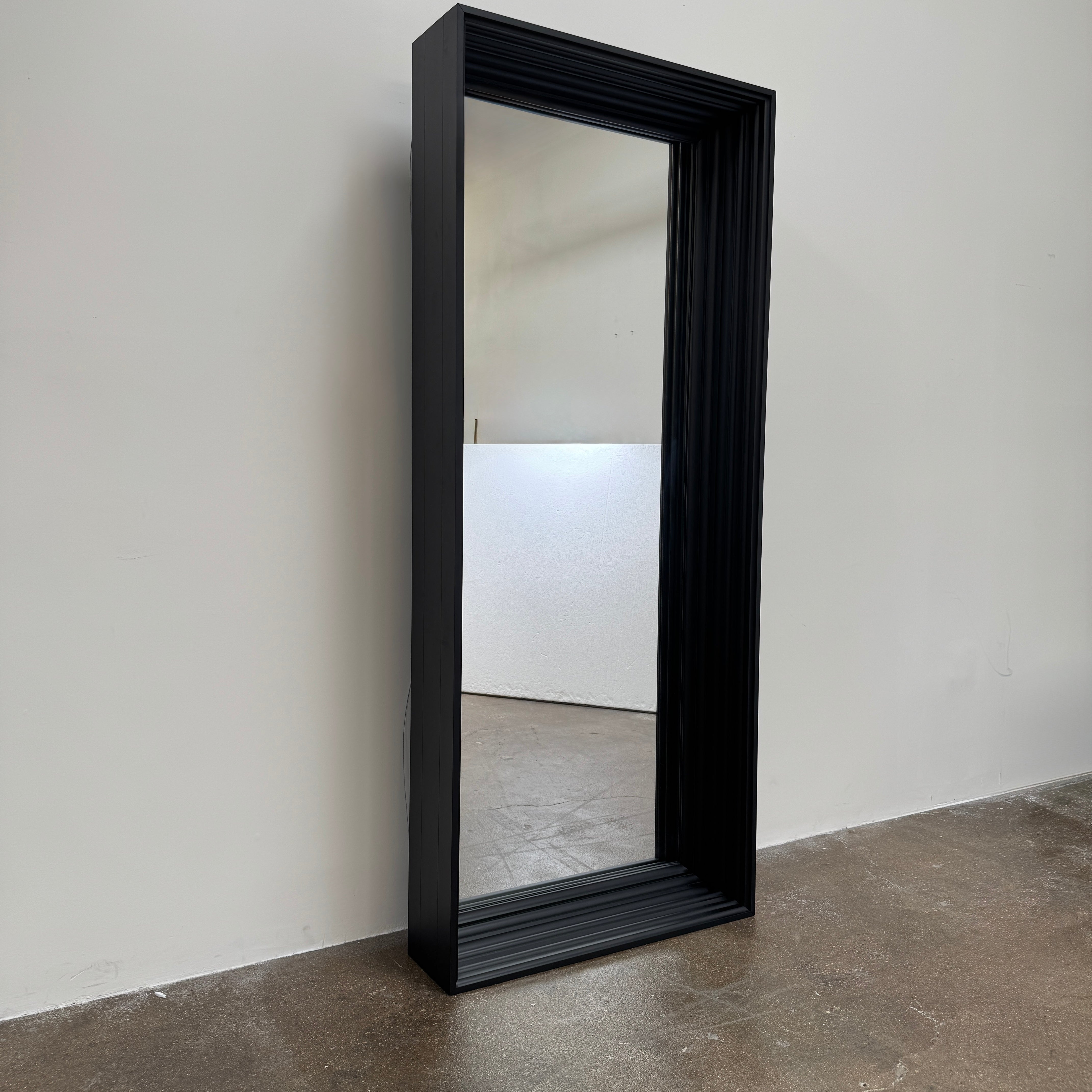 A person kneels in front of the Moooi Frame Mirror by Moooi—a large, rectangular mirror with a sleek black aluminum frame—while taking a photo. The mirror reflects furniture and industrial shelving within a well-lit room.