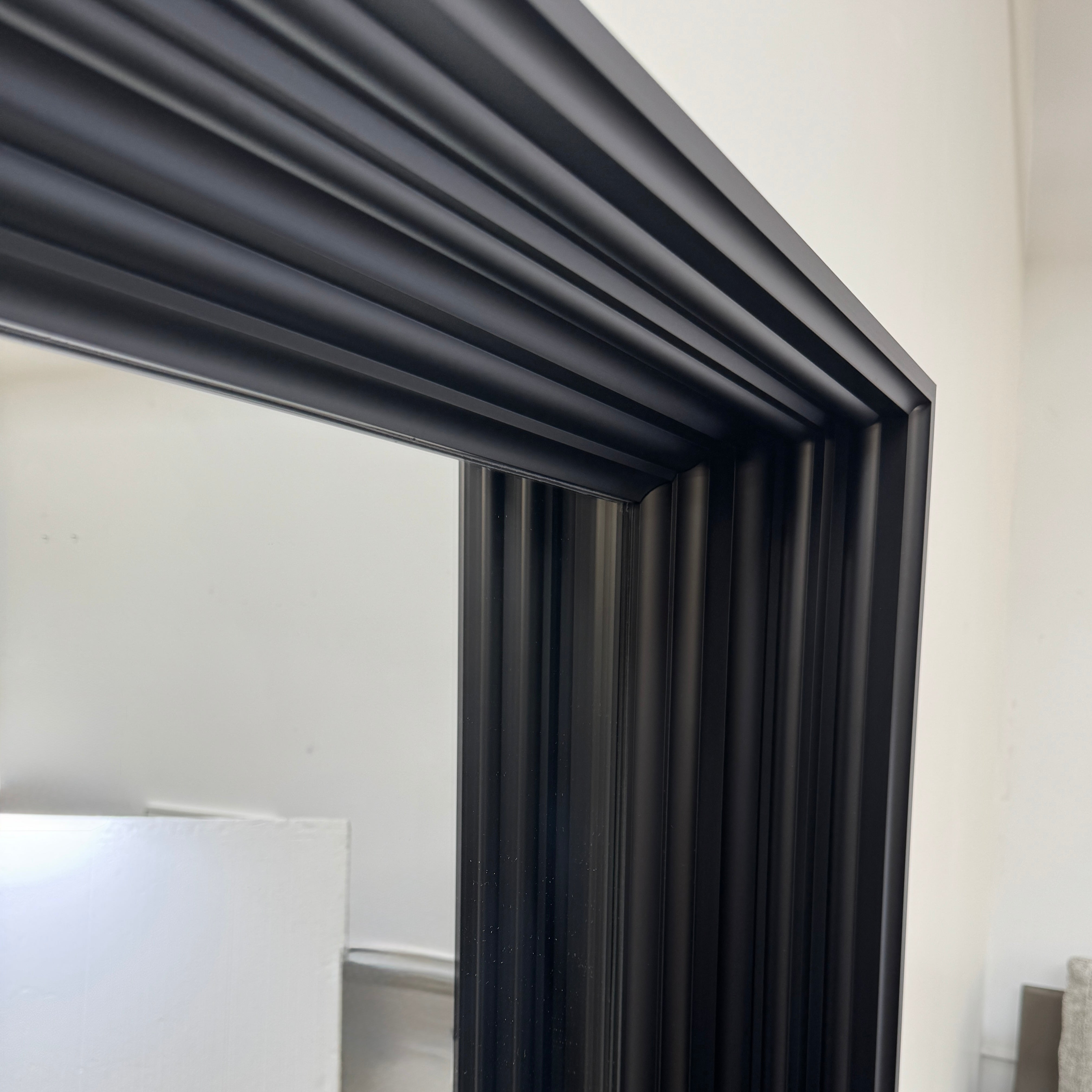 A person kneels in front of the Moooi Frame Mirror by Moooi—a large, rectangular mirror with a sleek black aluminum frame—while taking a photo. The mirror reflects furniture and industrial shelving within a well-lit room.
