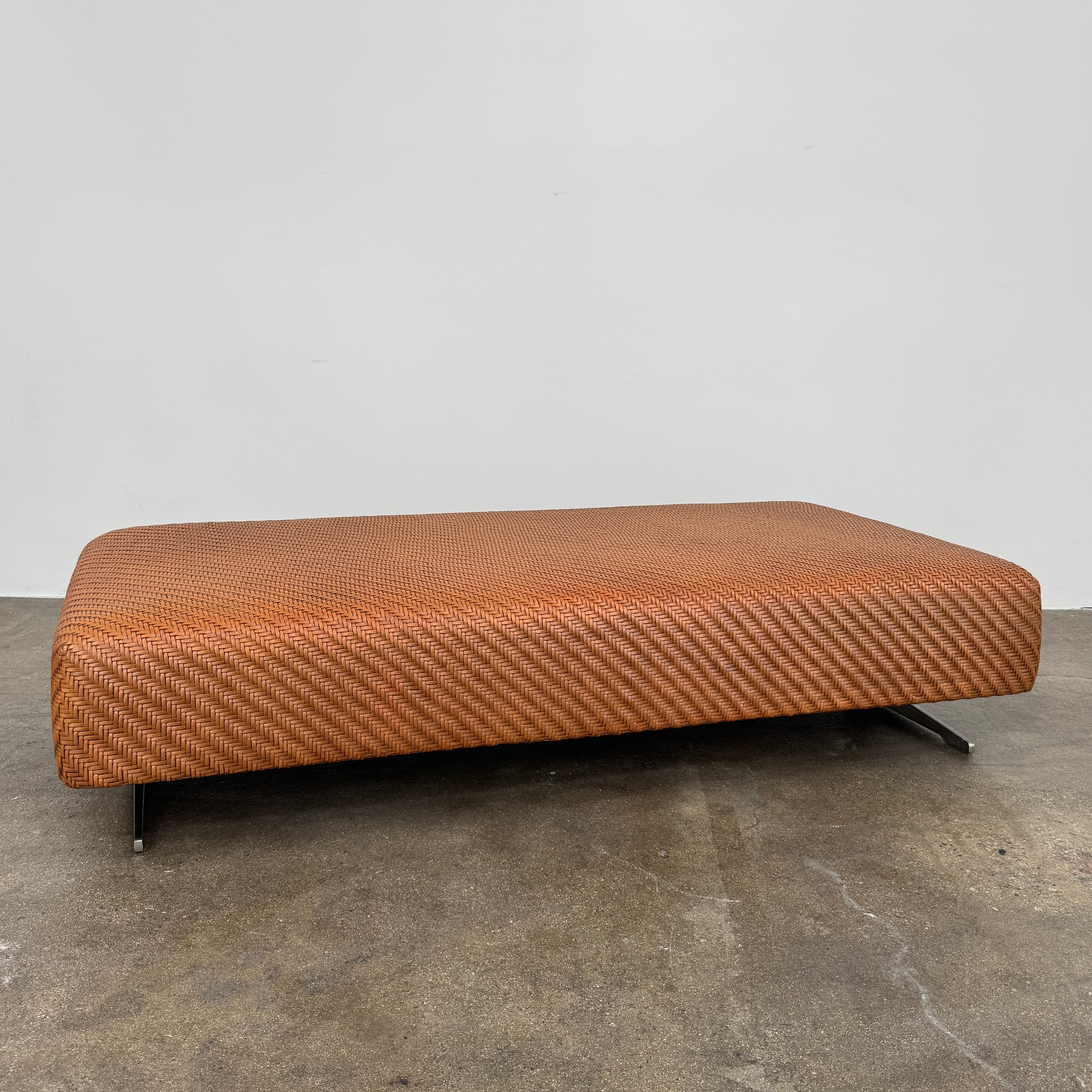 Flexform Filicudi Woven Pouf by Flexform, a rectangular orange bench with a low backless design and metal legs, displayed against a plain white background. Inspired by the iconic Filicudi collection, this elegant piece showcases a distinctive herringbone weave that adds texture and sophistication.