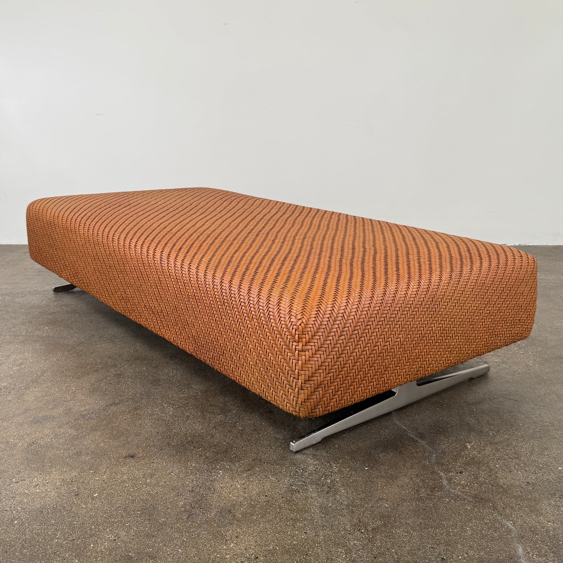 Flexform Filicudi Woven Pouf by Flexform, a rectangular orange bench with a low backless design and metal legs, displayed against a plain white background. Inspired by the iconic Filicudi collection, this elegant piece showcases a distinctive herringbone weave that adds texture and sophistication.