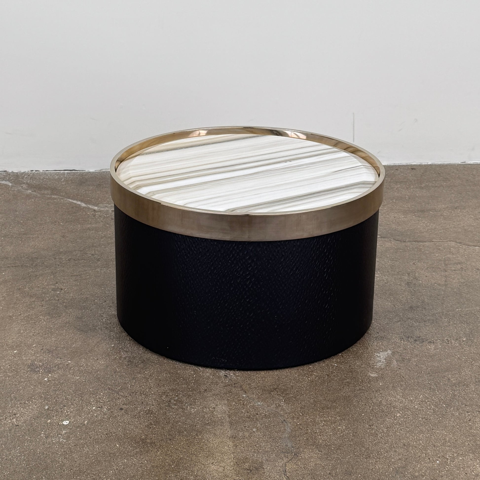 A round luxury accent table from Minotti, the Bally Accent Table features a black cylindrical base and a light-colored, striped marble top encircled by a slim metallic rim, elegantly displayed on a textured brown floor. Inspired by refined design, this signature piece from Minotti makes an essential addition to any luxury furniture collection.