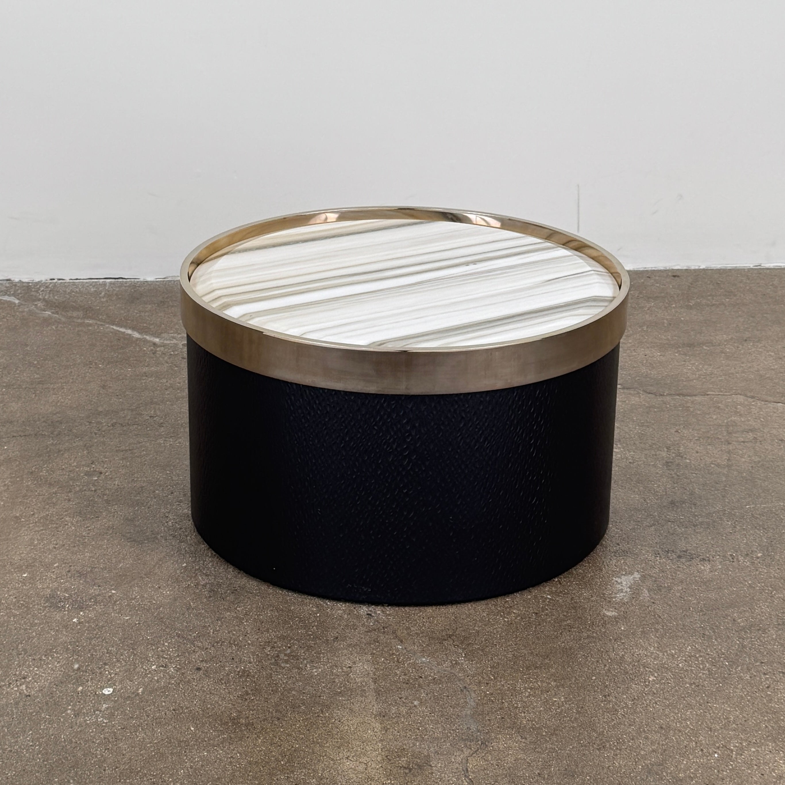 A round luxury accent table from Minotti, the Bally Accent Table features a black cylindrical base and a light-colored, striped marble top encircled by a slim metallic rim, elegantly displayed on a textured brown floor. Inspired by refined design, this signature piece from Minotti makes an essential addition to any luxury furniture collection.