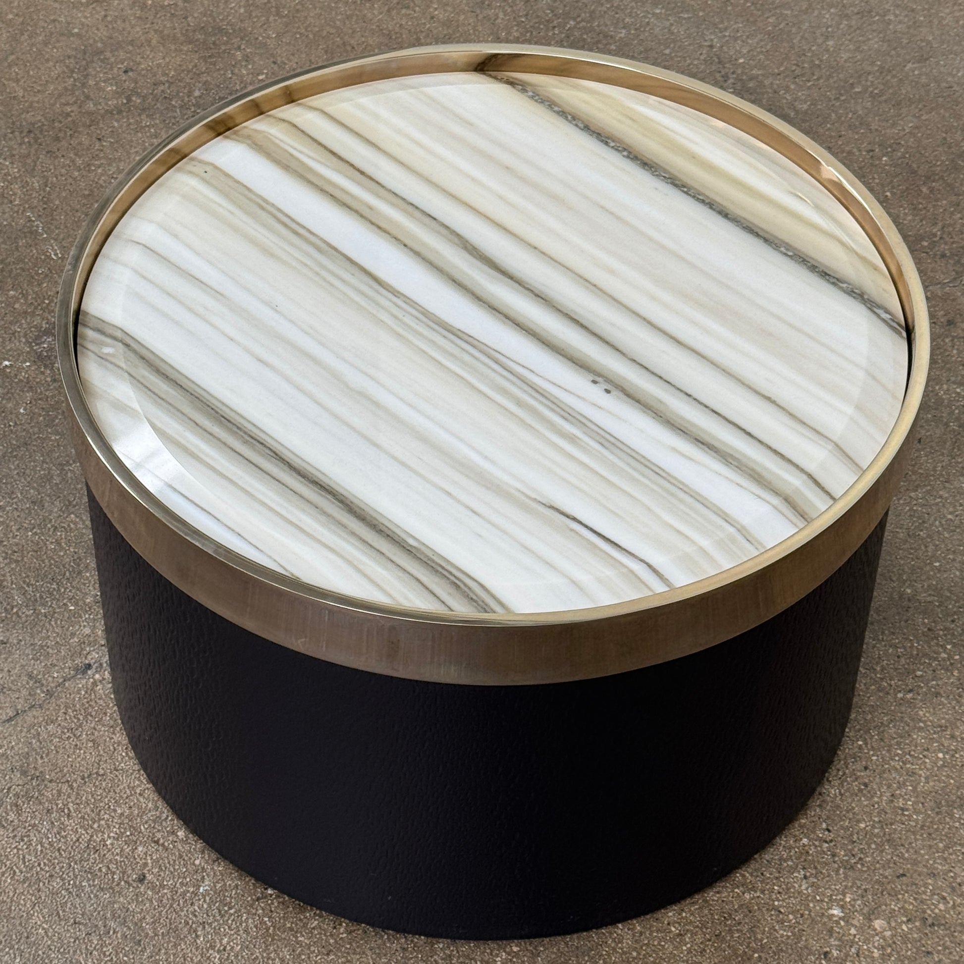 A round luxury accent table from Minotti, the Bally Accent Table features a black cylindrical base and a light-colored, striped marble top encircled by a slim metallic rim, elegantly displayed on a textured brown floor. Inspired by refined design, this signature piece from Minotti makes an essential addition to any luxury furniture collection.