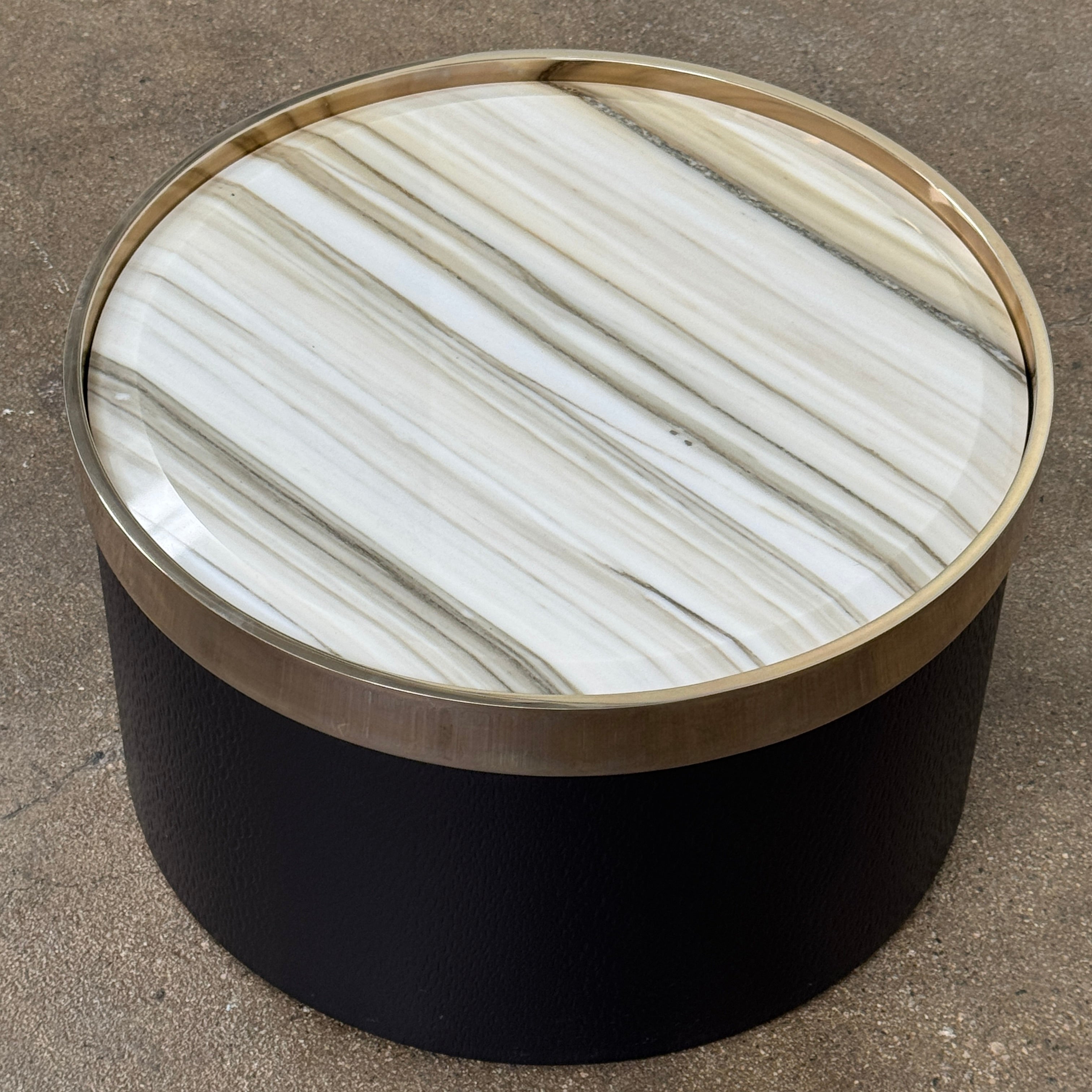 A round luxury accent table from Minotti, the Bally Accent Table features a black cylindrical base and a light-colored, striped marble top encircled by a slim metallic rim, elegantly displayed on a textured brown floor. Inspired by refined design, this signature piece from Minotti makes an essential addition to any luxury furniture collection.