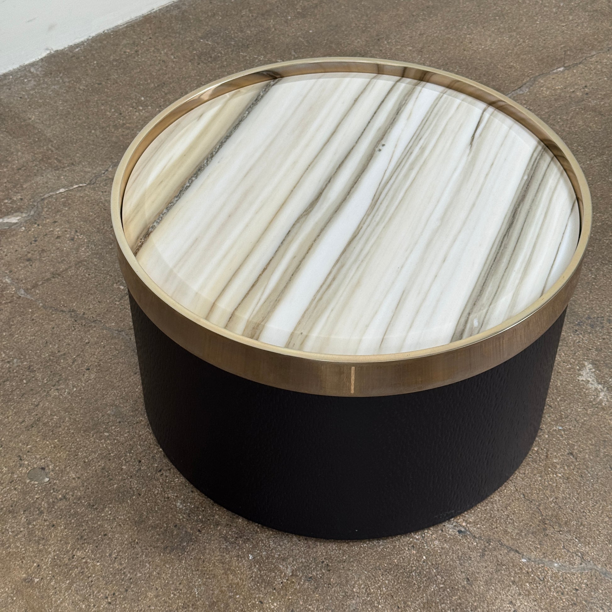 A round luxury accent table from Minotti, the Bally Accent Table features a black cylindrical base and a light-colored, striped marble top encircled by a slim metallic rim, elegantly displayed on a textured brown floor. Inspired by refined design, this signature piece from Minotti makes an essential addition to any luxury furniture collection.