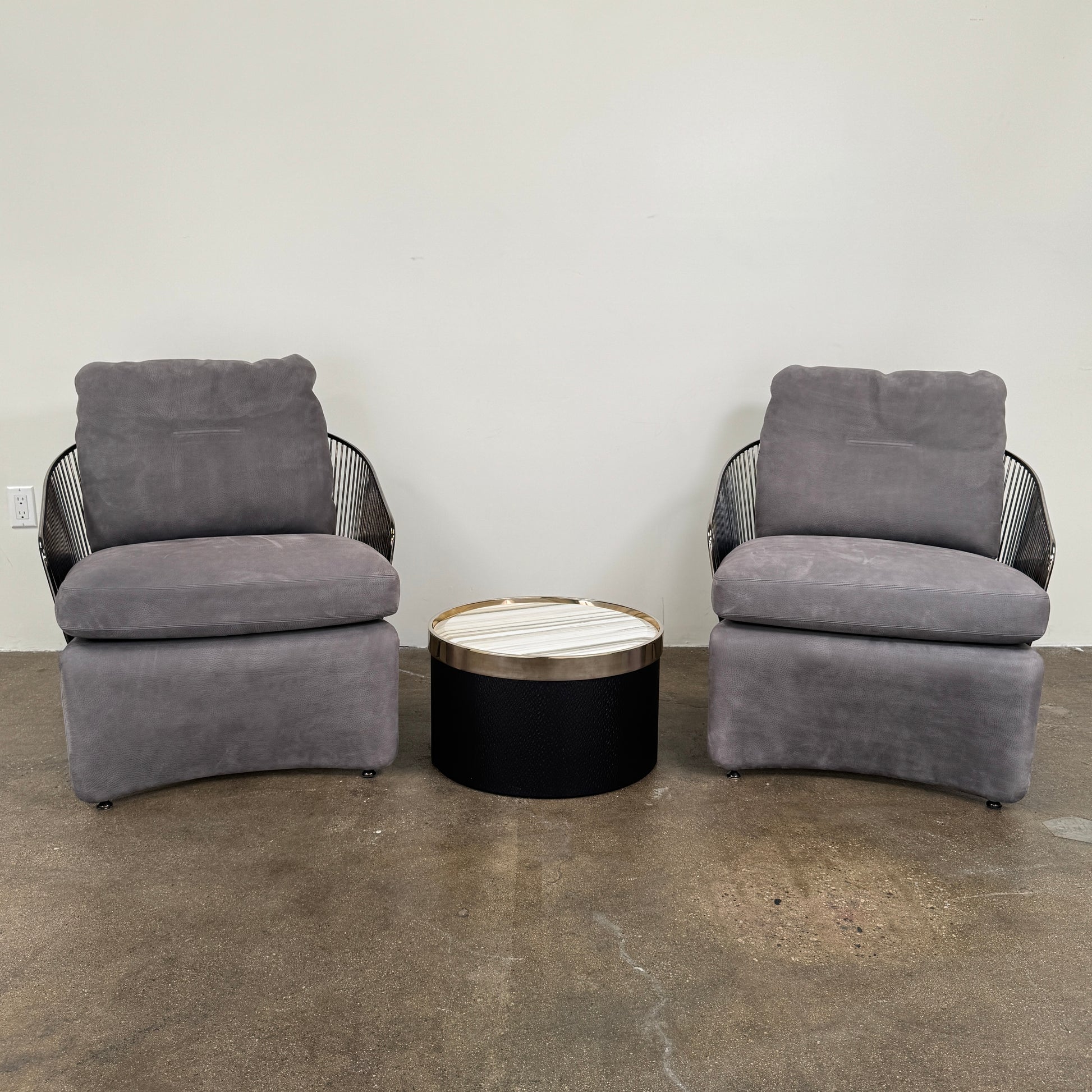 A set of two Minotti Colette Lounge Chairs by Minotti is displayed side by side against a plain white background. Each chair showcases luxurious gray nubuck leather upholstery and sleek black metal armrests, elegantly capturing the timeless mid-century silhouette characteristic of the iconic design.