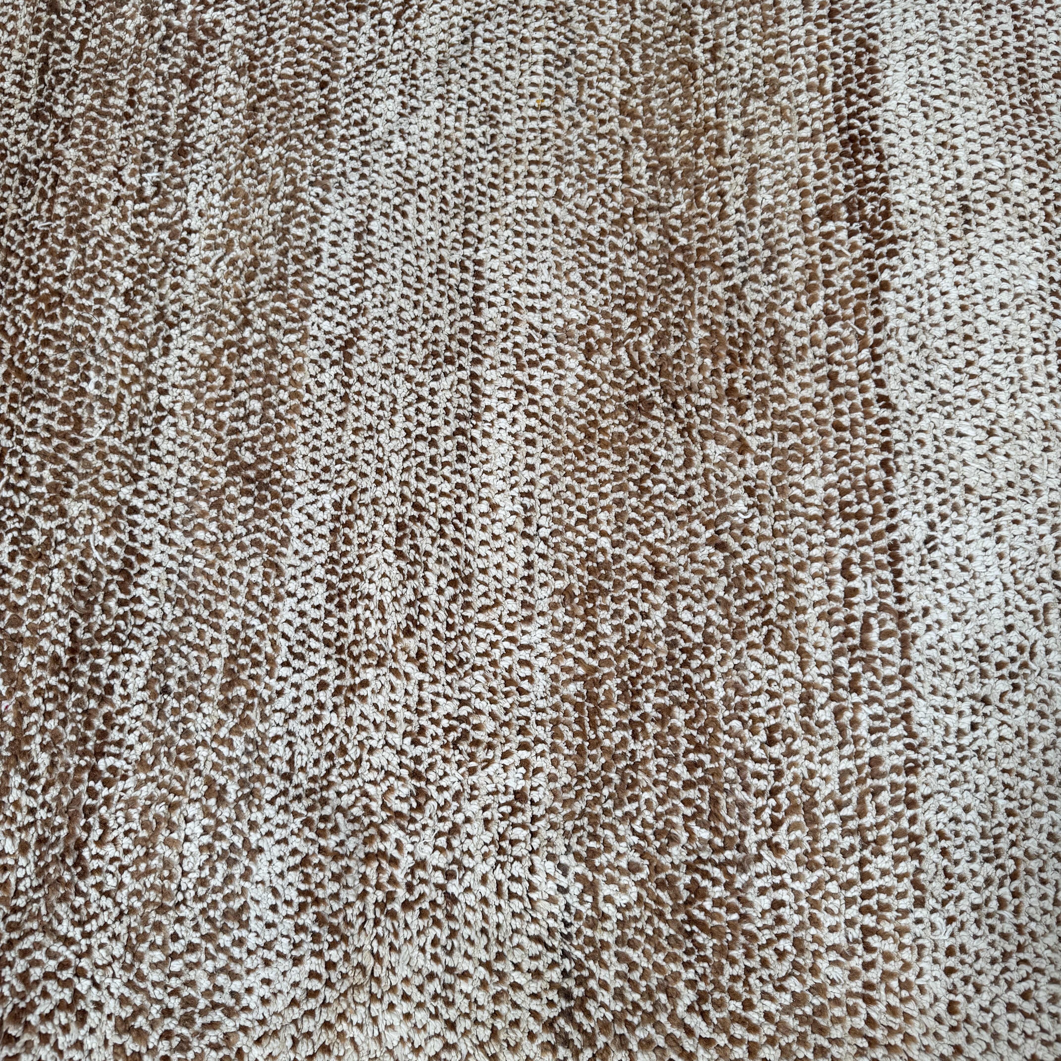 Close-up of the ABC Rugs Brown Solid Textured Silk Rug (8' x 10') from ABC Carpet and Home, featuring beige and brown tones with a subtly mottled texture. A small fabric tag appears in the top left corner.