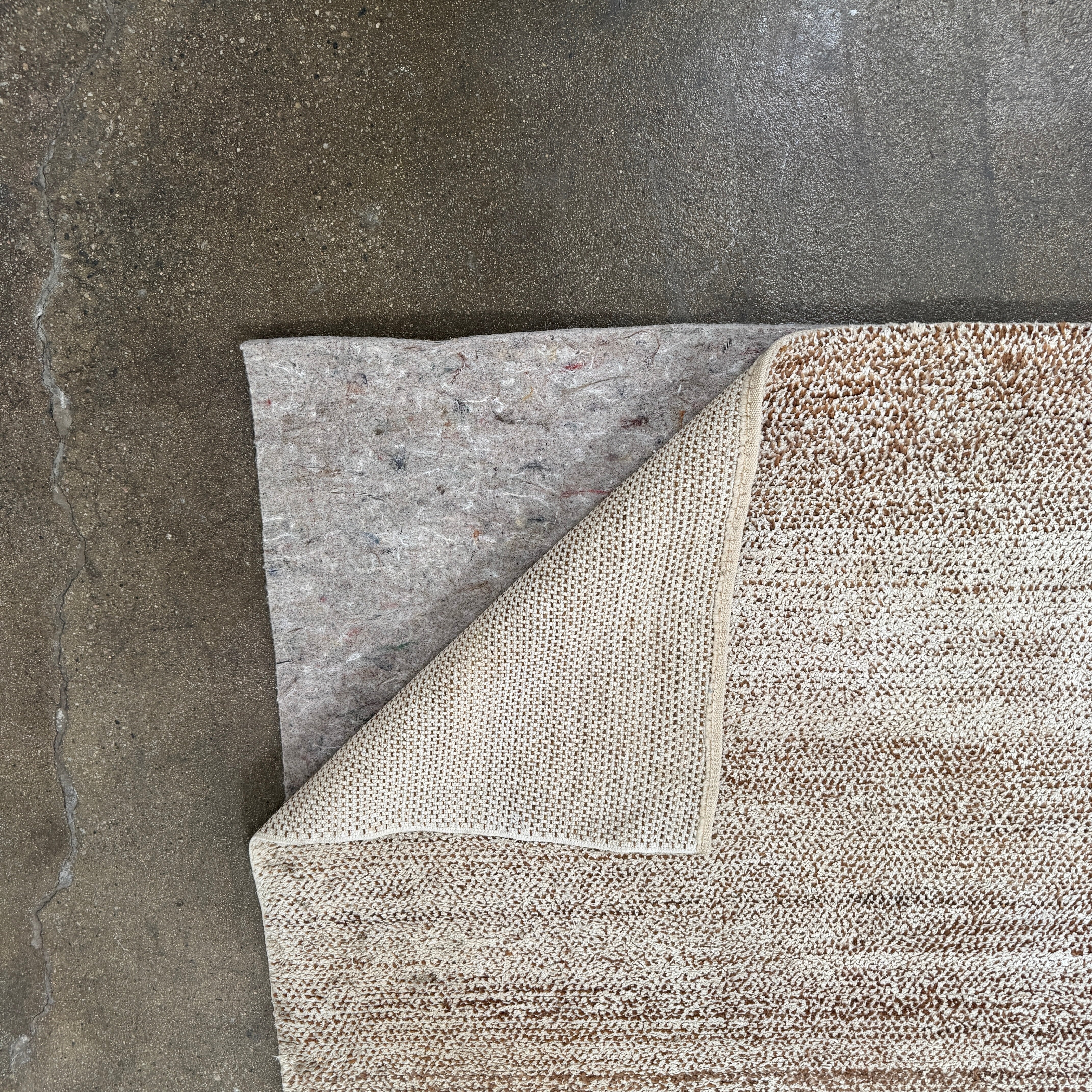 Close-up of the ABC Rugs Brown Solid Textured Silk Rug (8' x 10') from ABC Carpet and Home, featuring beige and brown tones with a subtly mottled texture. A small fabric tag appears in the top left corner.