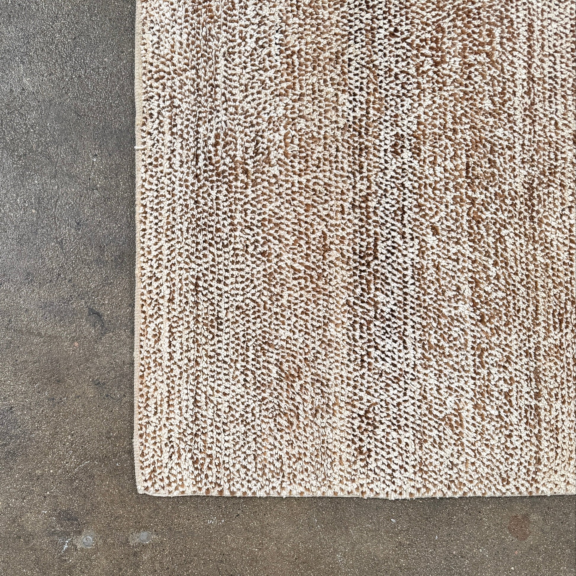 Close-up of the ABC Rugs Brown Solid Textured Silk Rug (8' x 10') from ABC Carpet and Home, featuring beige and brown tones with a subtly mottled texture. A small fabric tag appears in the top left corner.