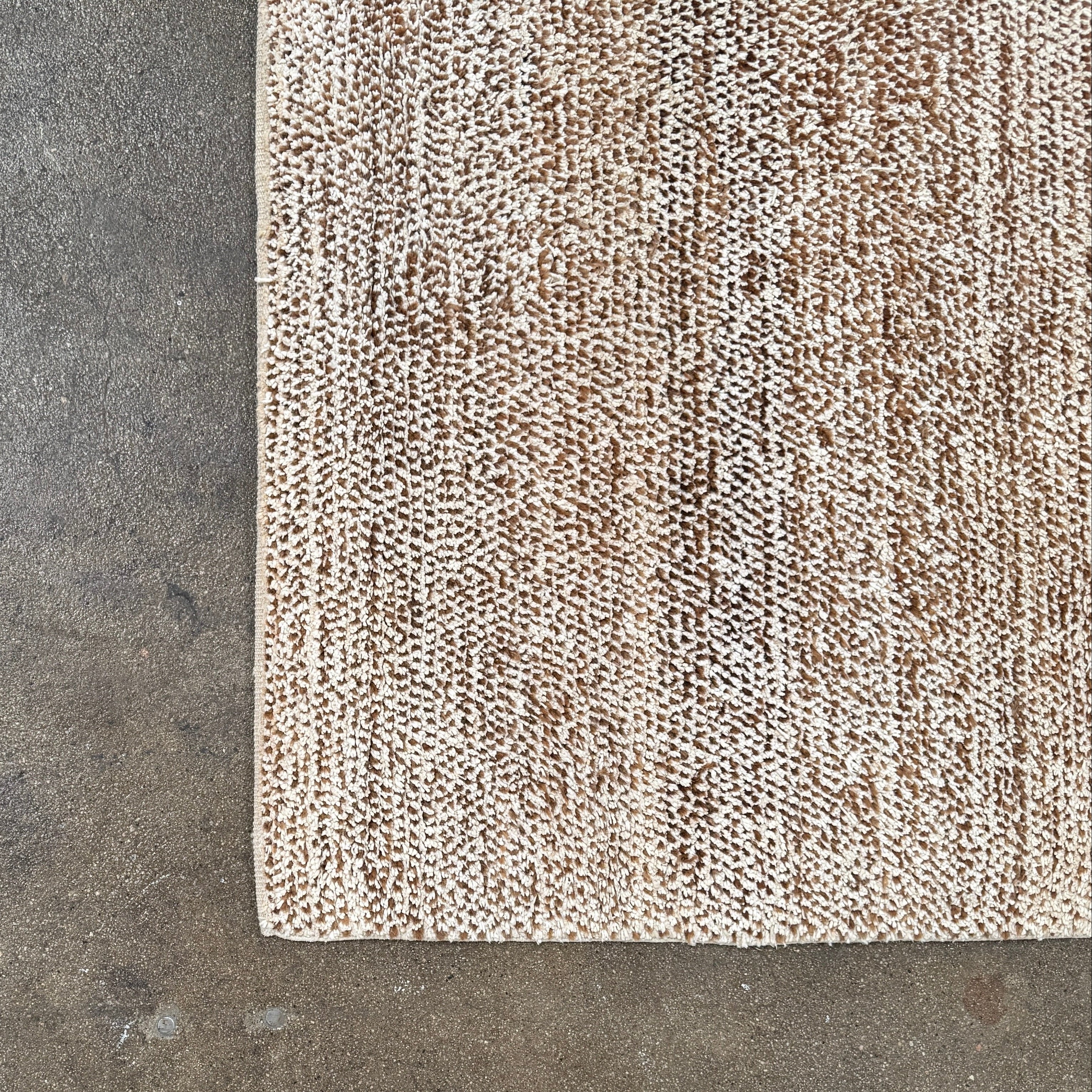 Close-up of the ABC Rugs Brown Solid Textured Silk Rug (8' x 10') from ABC Carpet and Home, featuring beige and brown tones with a subtly mottled texture. A small fabric tag appears in the top left corner.