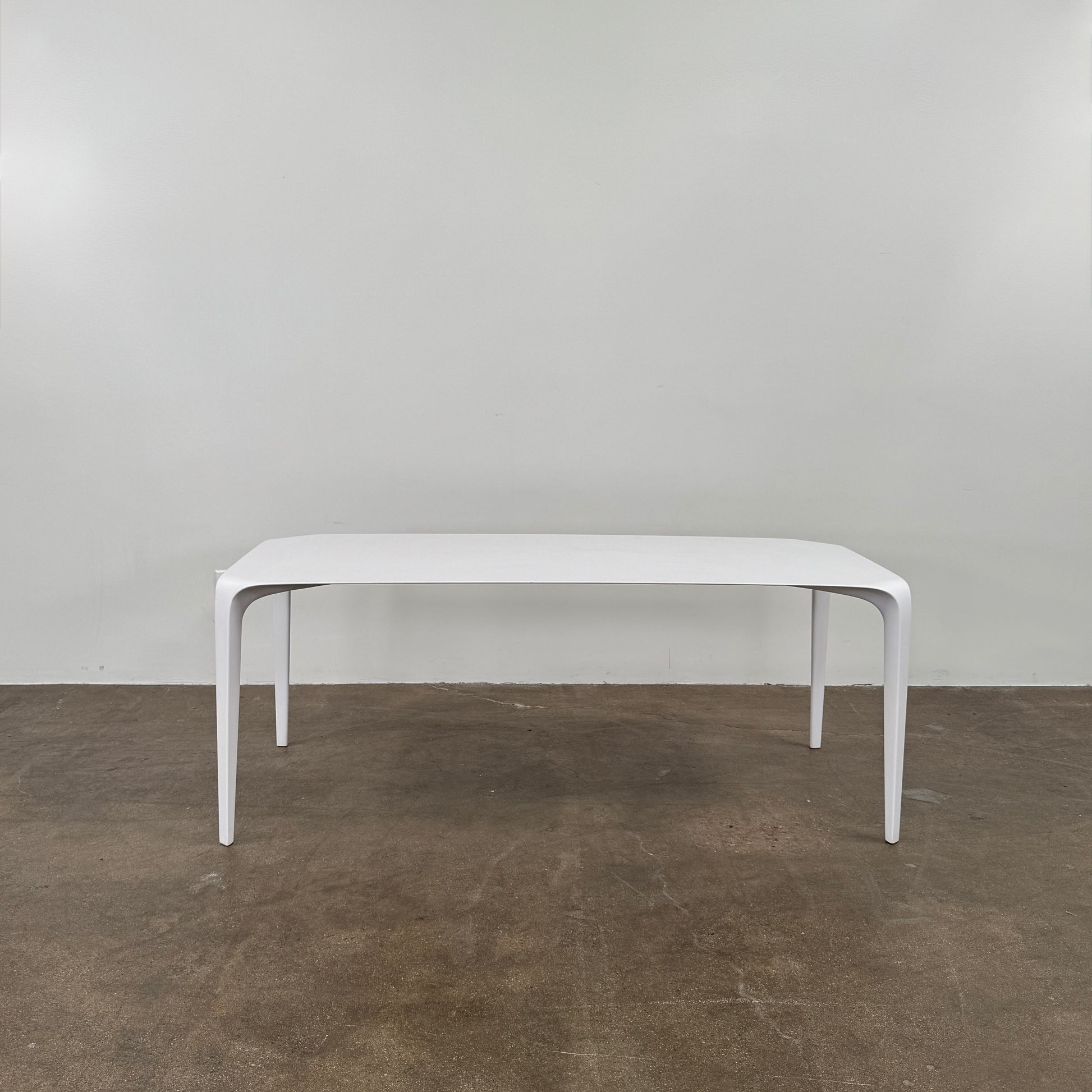 A modern white rectangular B&B Italia LINK Indoor / Outdoor Dining Table by B&B Italia, featuring rounded edges, stands on a brown concrete floor against a plain light gray wall.