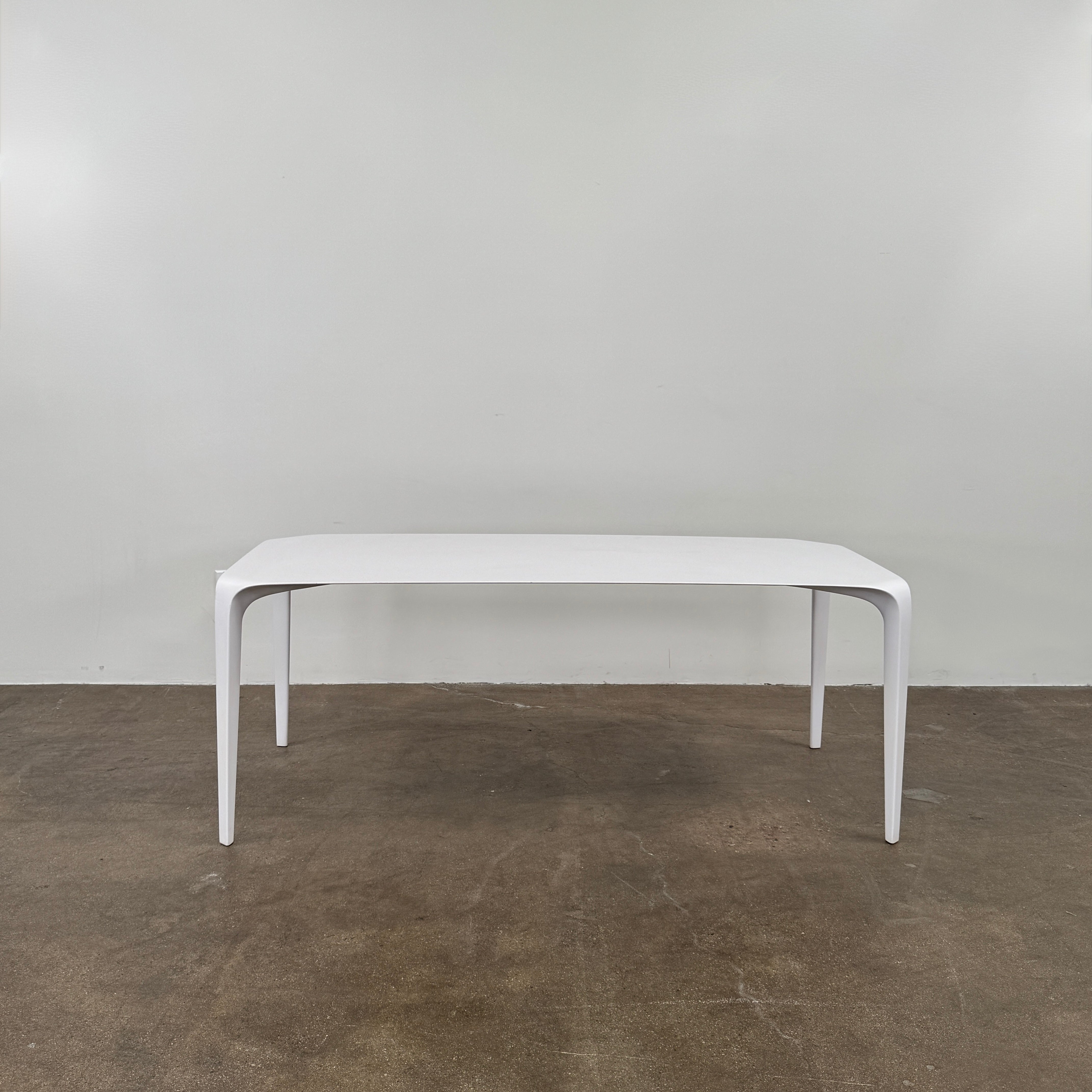 A modern white rectangular B&B Italia LINK Indoor / Outdoor Dining Table by B&B Italia, featuring rounded edges, stands on a brown concrete floor against a plain light gray wall.