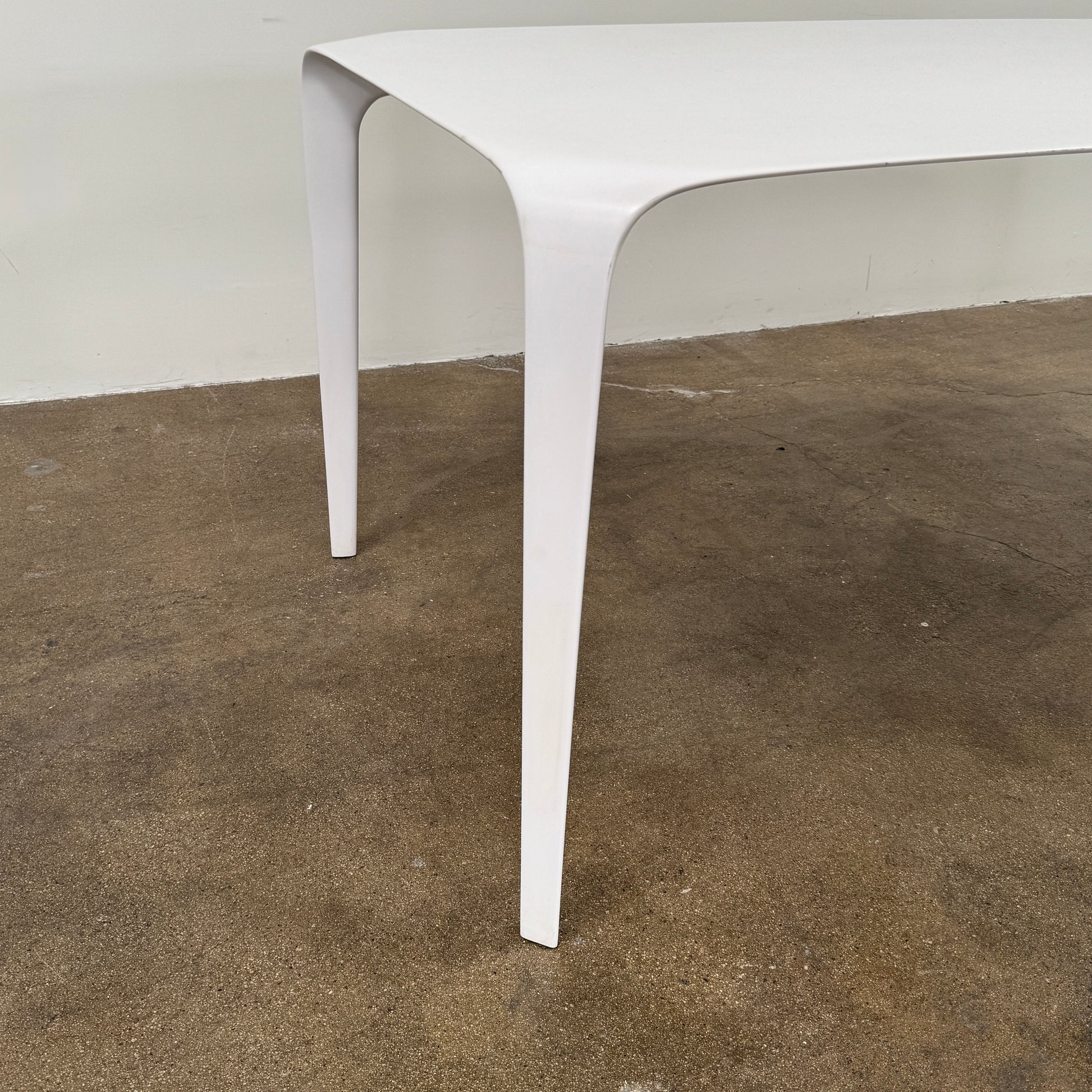 A modern white rectangular B&B Italia LINK Indoor / Outdoor Dining Table by B&B Italia, featuring rounded edges, stands on a brown concrete floor against a plain light gray wall.