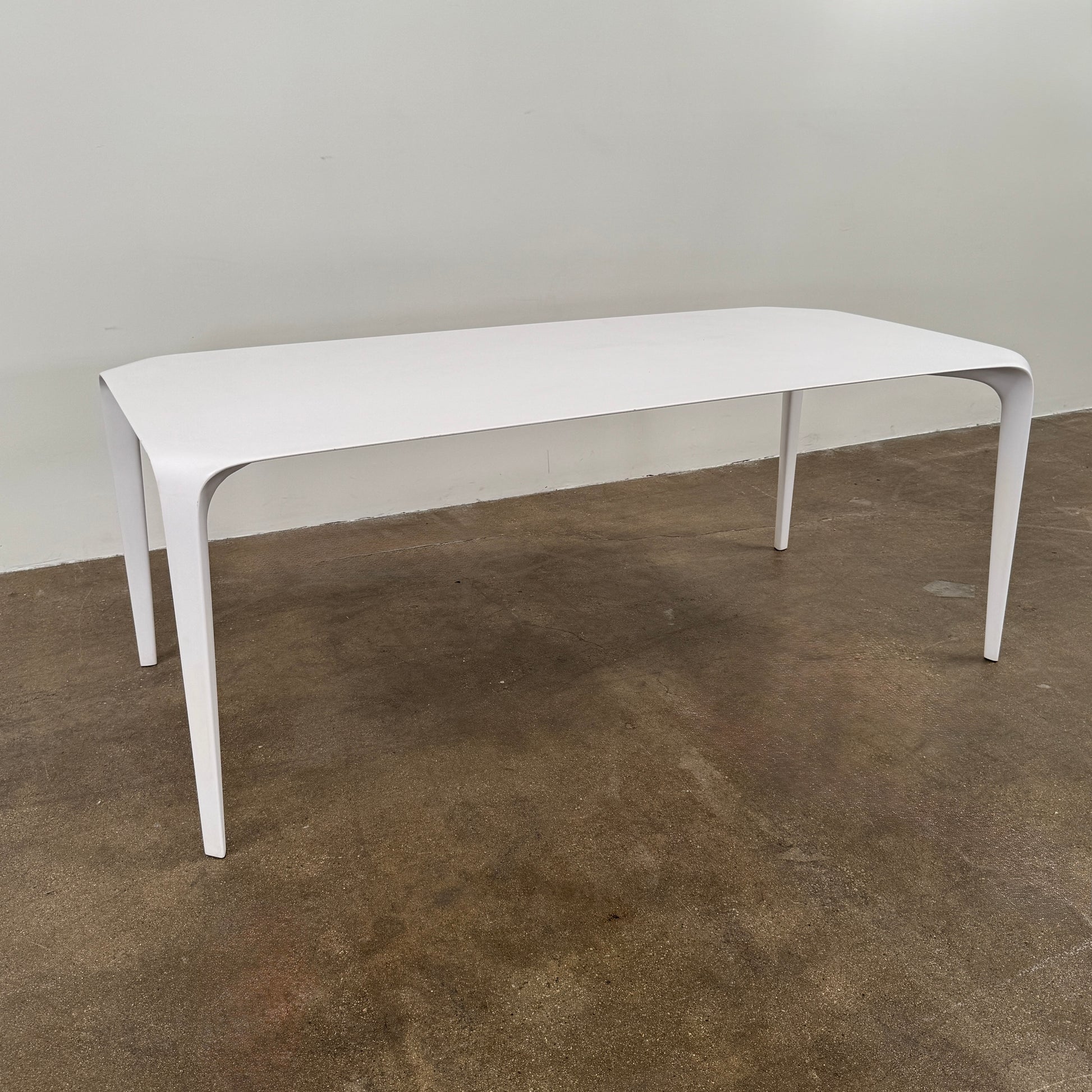 A modern white rectangular B&B Italia LINK Indoor / Outdoor Dining Table by B&B Italia, featuring rounded edges, stands on a brown concrete floor against a plain light gray wall.