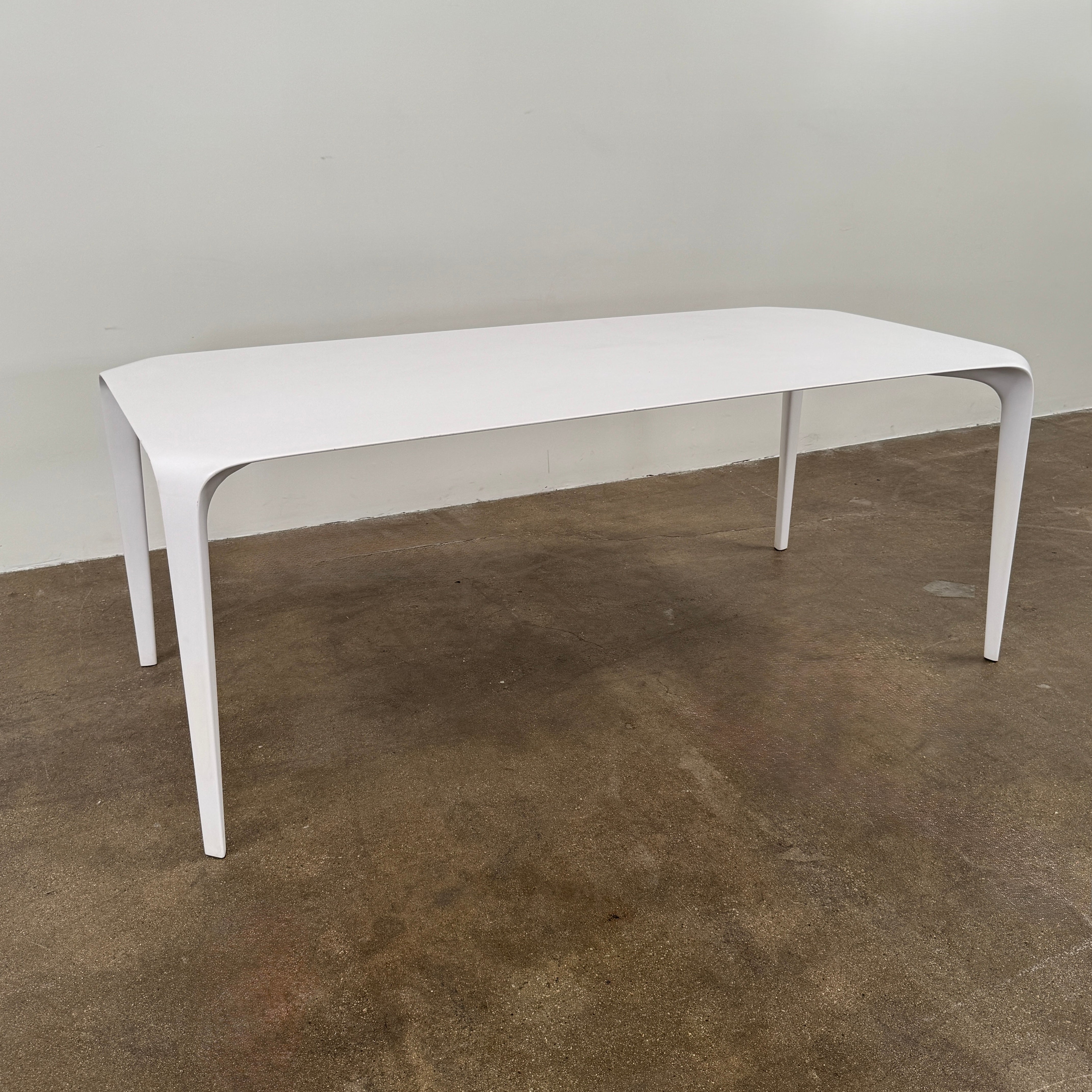 A modern white rectangular B&B Italia LINK Indoor / Outdoor Dining Table by B&B Italia, featuring rounded edges, stands on a brown concrete floor against a plain light gray wall.