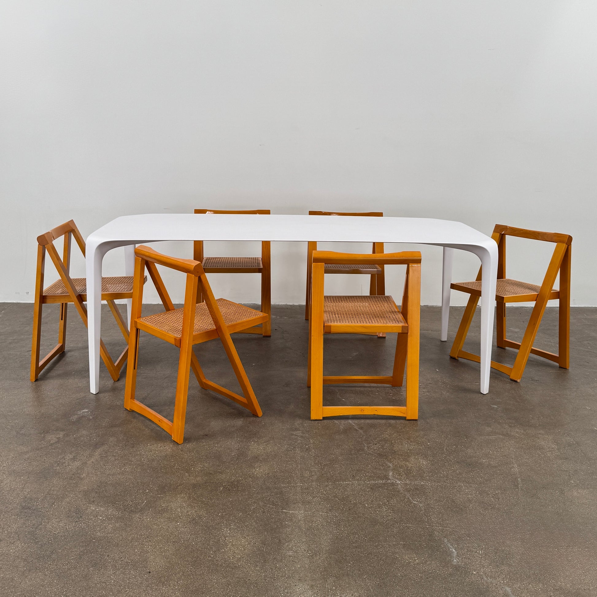 A set of 2 Vintage Aldo Jacober Folding Chairs, featuring wooden frames and woven rattan seats, are positioned side by side on a concrete floor against a plain white wall, shown from the back.