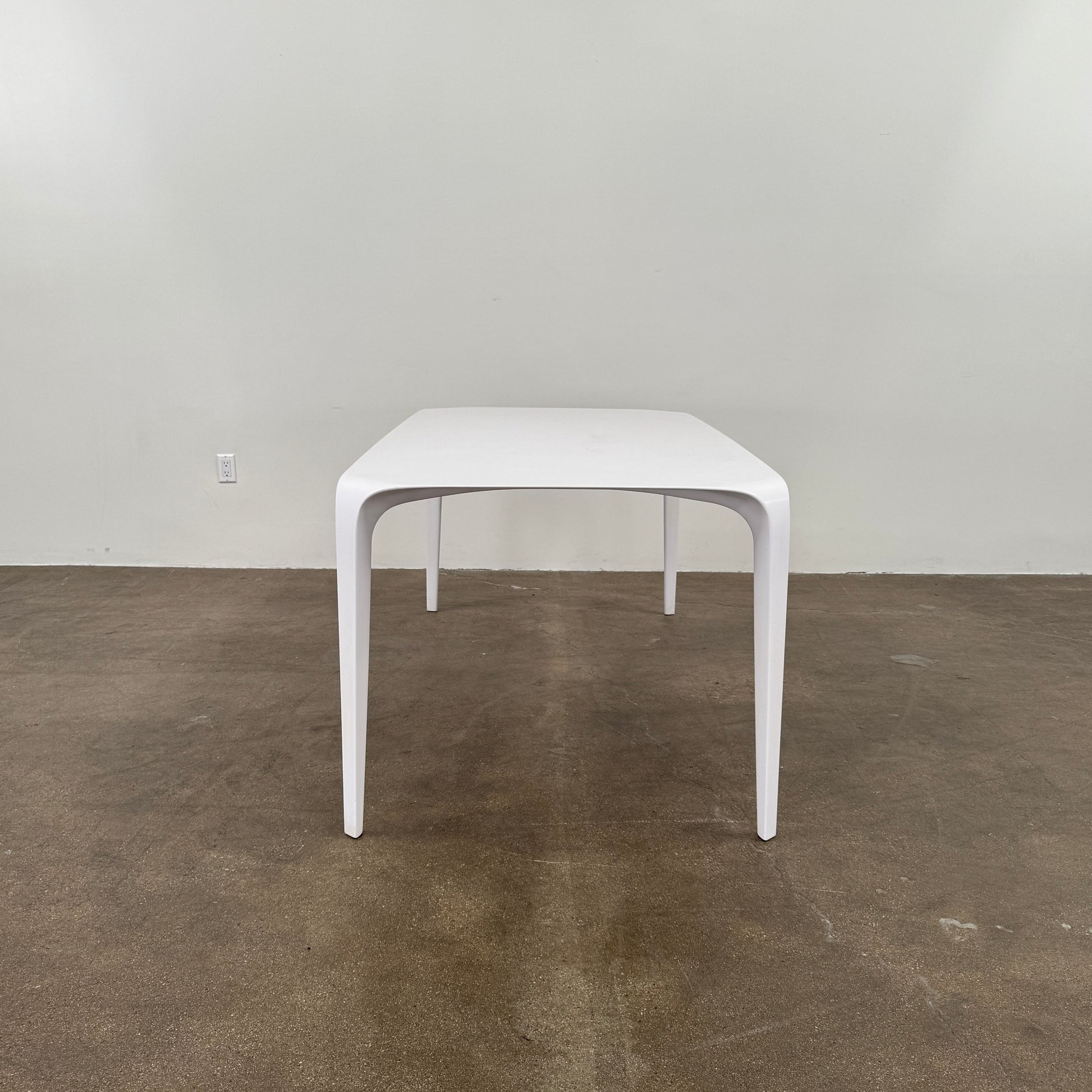 A modern white rectangular B&B Italia LINK Indoor / Outdoor Dining Table by B&B Italia, featuring rounded edges, stands on a brown concrete floor against a plain light gray wall.