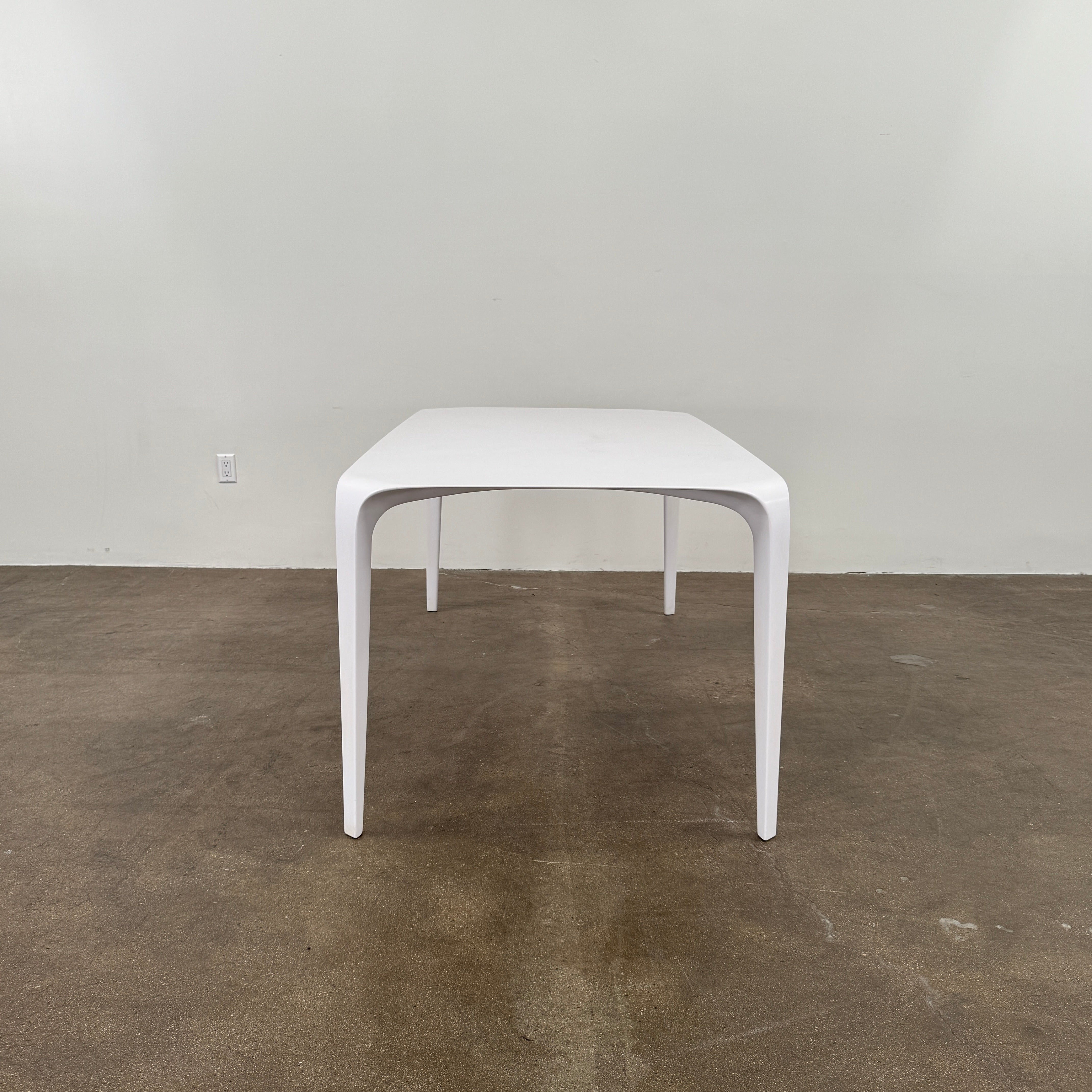 A modern white rectangular B&B Italia LINK Indoor / Outdoor Dining Table by B&B Italia, featuring rounded edges, stands on a brown concrete floor against a plain light gray wall.