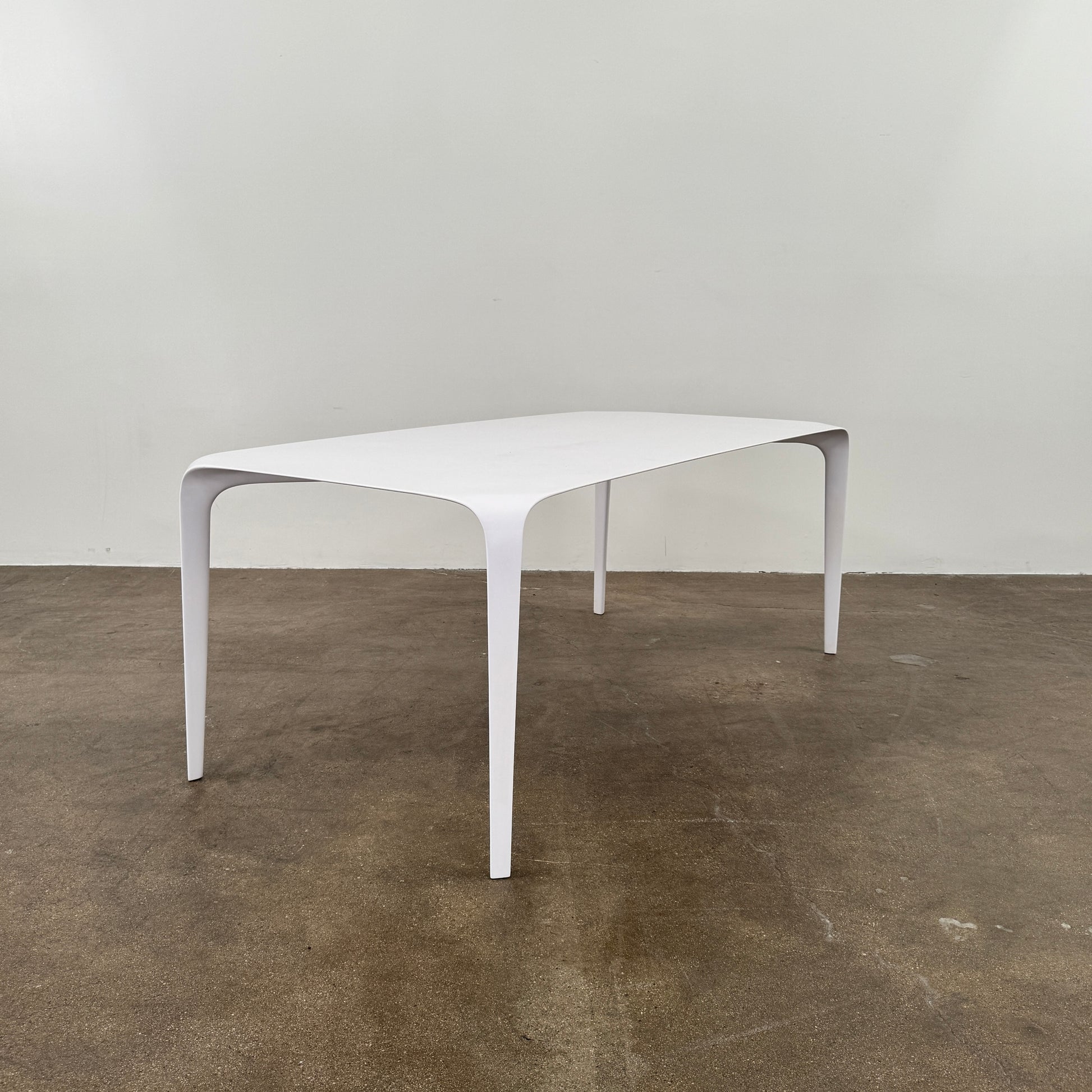 A modern white rectangular B&B Italia LINK Indoor / Outdoor Dining Table by B&B Italia, featuring rounded edges, stands on a brown concrete floor against a plain light gray wall.