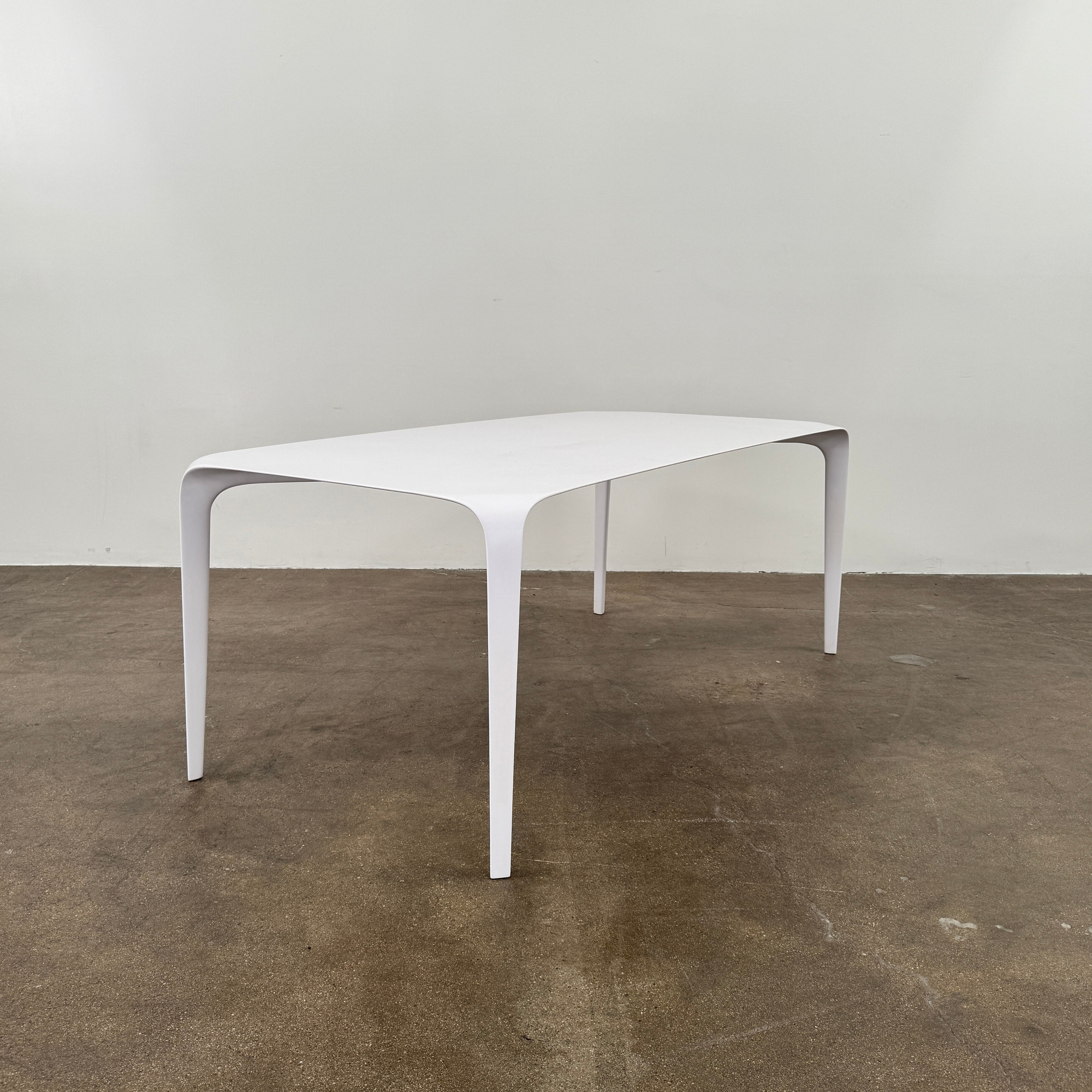 A modern white rectangular B&B Italia LINK Indoor / Outdoor Dining Table by B&B Italia, featuring rounded edges, stands on a brown concrete floor against a plain light gray wall.