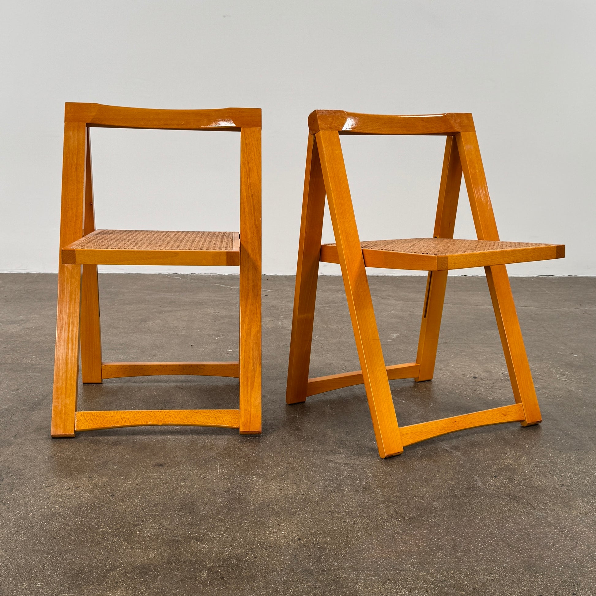 A set of 2 Vintage Aldo Jacober Folding Chairs, featuring wooden frames and woven rattan seats, are positioned side by side on a concrete floor against a plain white wall, shown from the back.