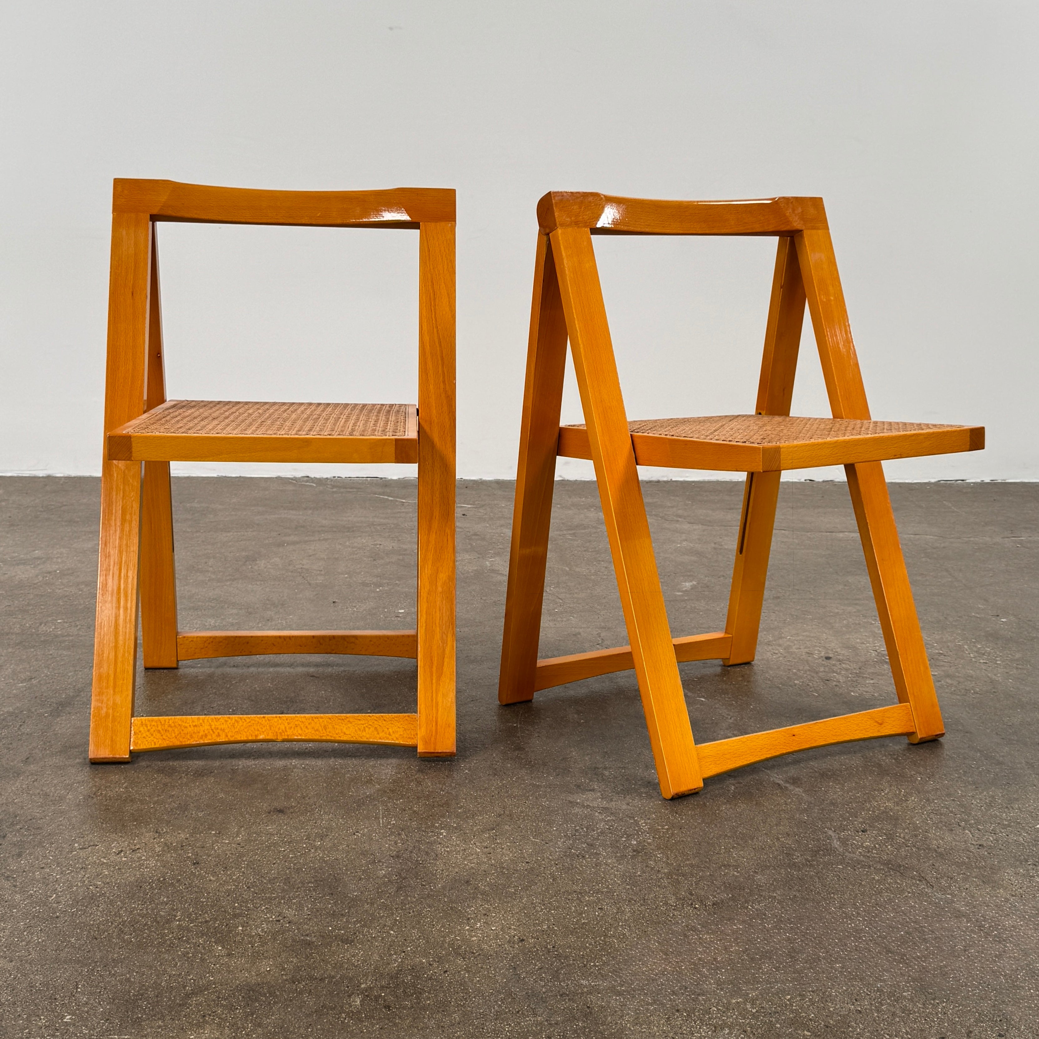 A set of 2 Vintage Aldo Jacober Folding Chairs, featuring wooden frames and woven rattan seats, are positioned side by side on a concrete floor against a plain white wall, shown from the back.