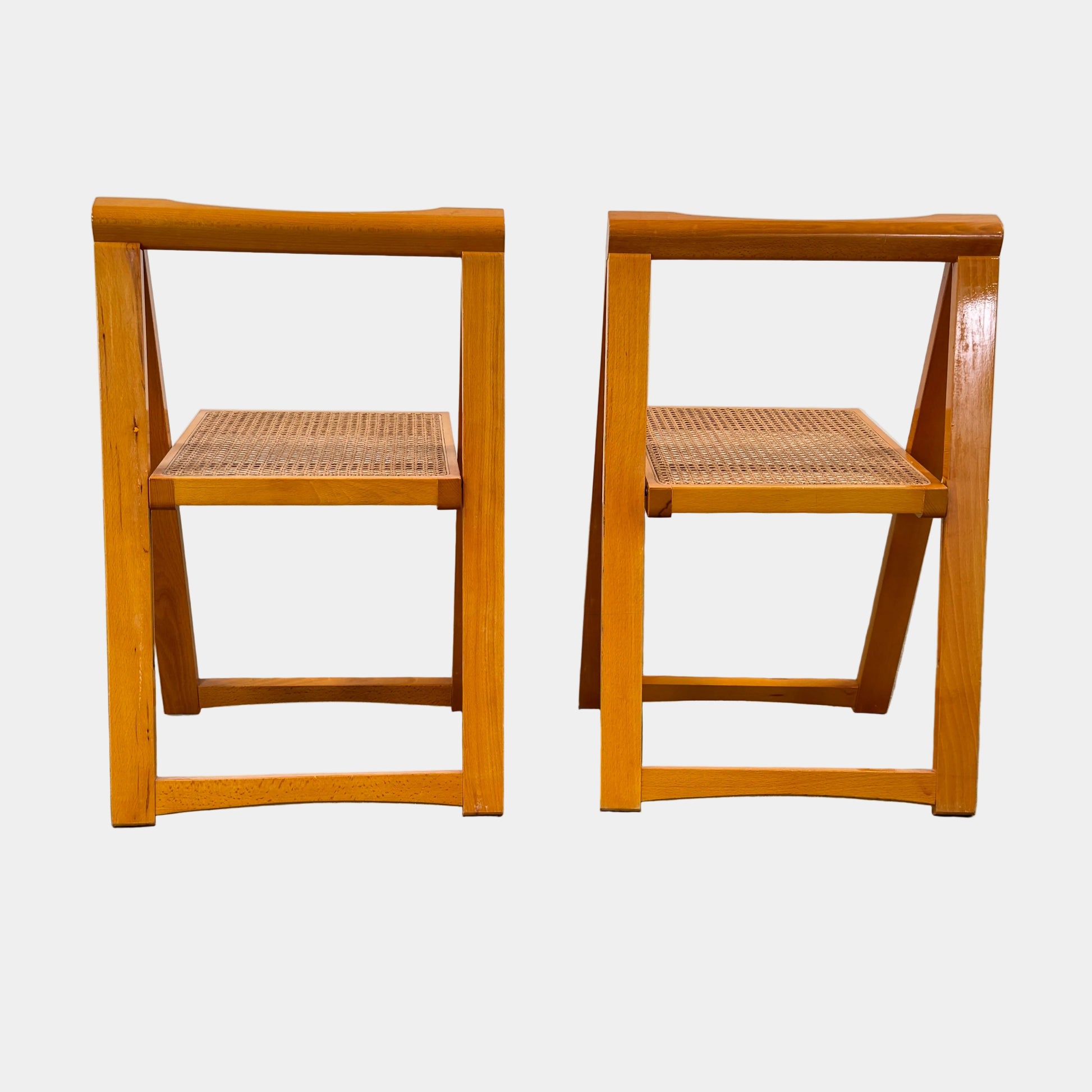 A set of 2 Vintage Aldo Jacober Folding Chairs, featuring wooden frames and woven rattan seats, are positioned side by side on a concrete floor against a plain white wall, shown from the back.
