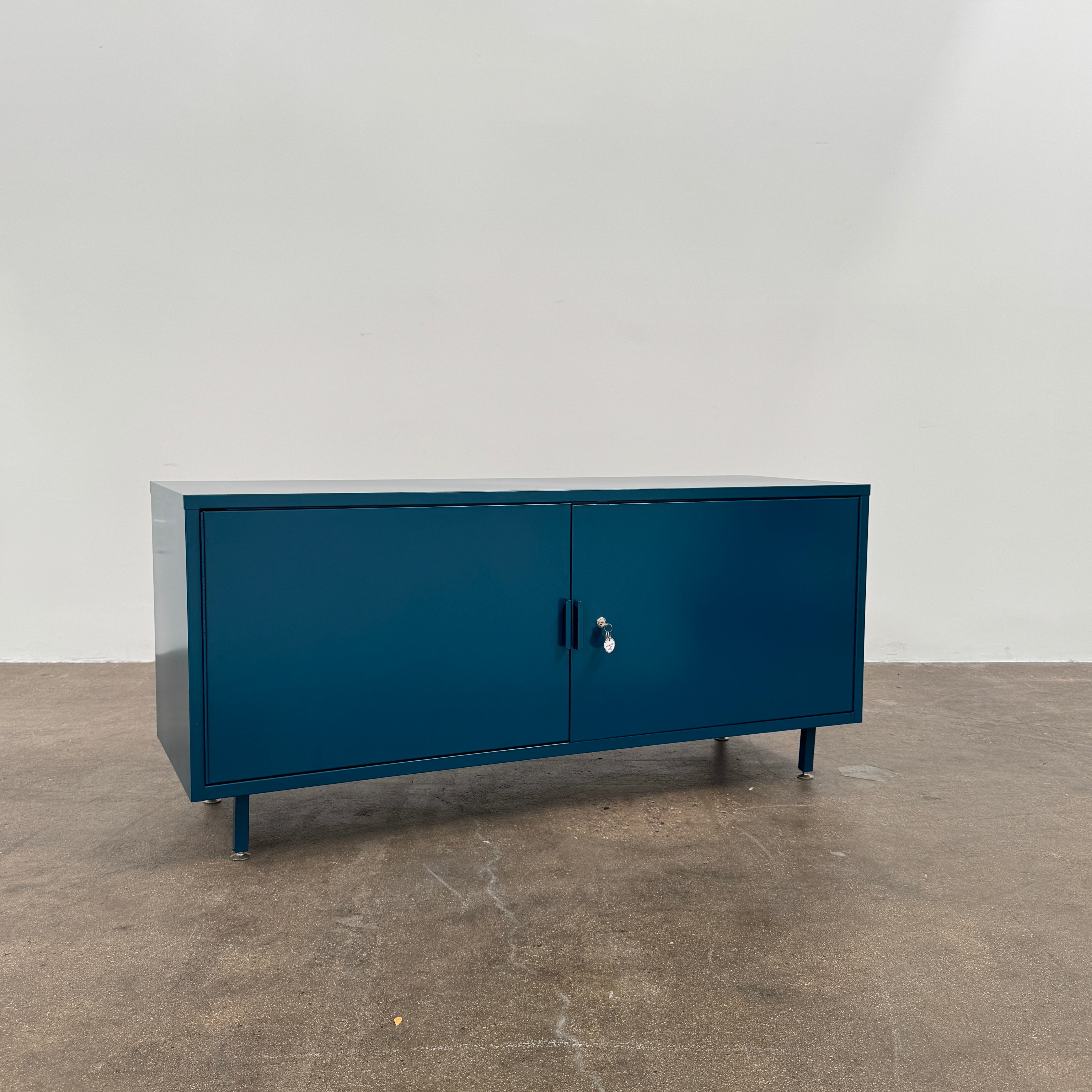 A blue metal Heartwork Active Duty Storage Credenza with two doors, a central lock, and four legs stands against a plain white background—this Heartwork piece offers durable construction ideal for workplace storage.