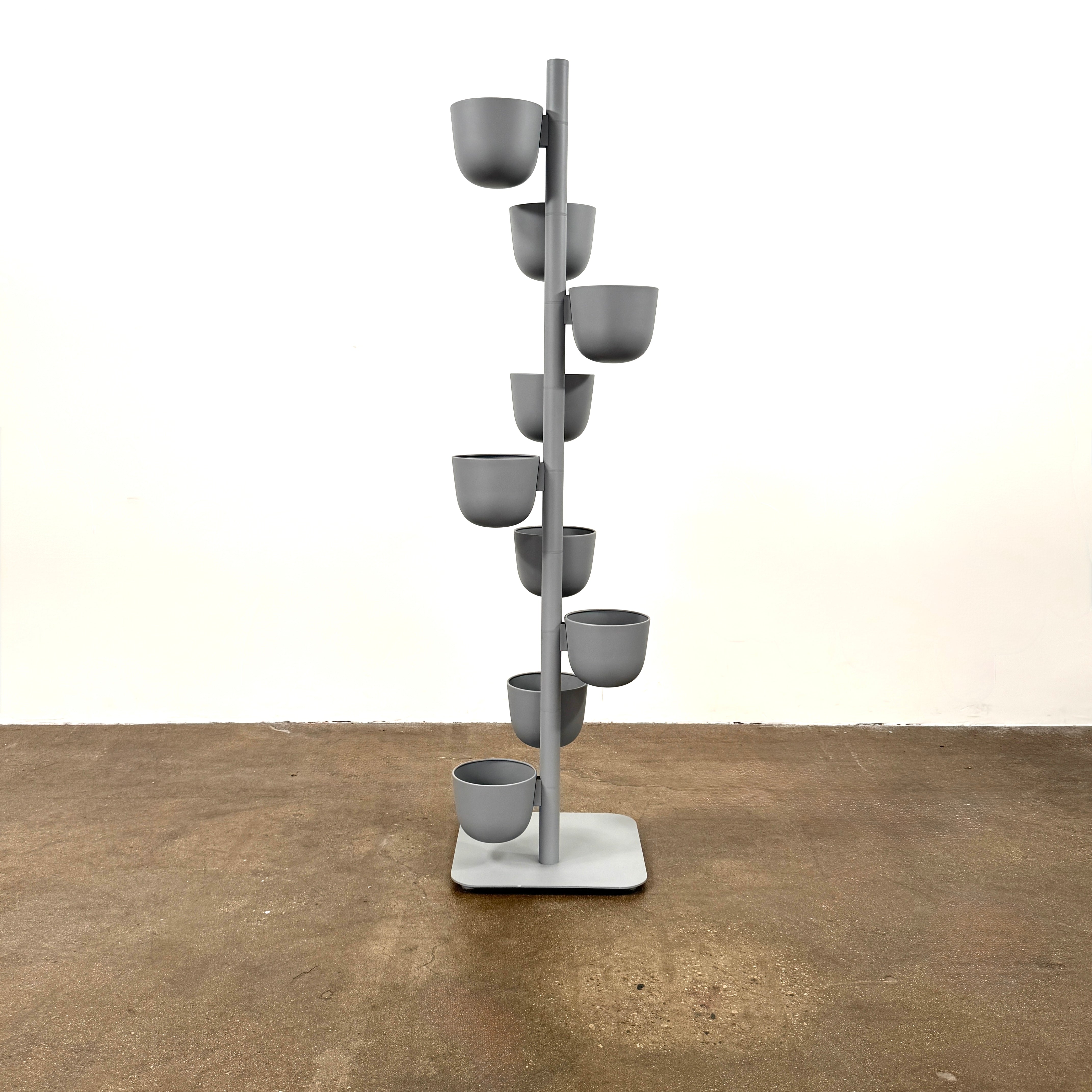 The DWR Story Planter by DWR is a vertical metal plant stand featuring eight gray round pots arranged in a staggered pattern along a single pole with a square base, making it an ideal decorative planter for fans of vertical gardening.