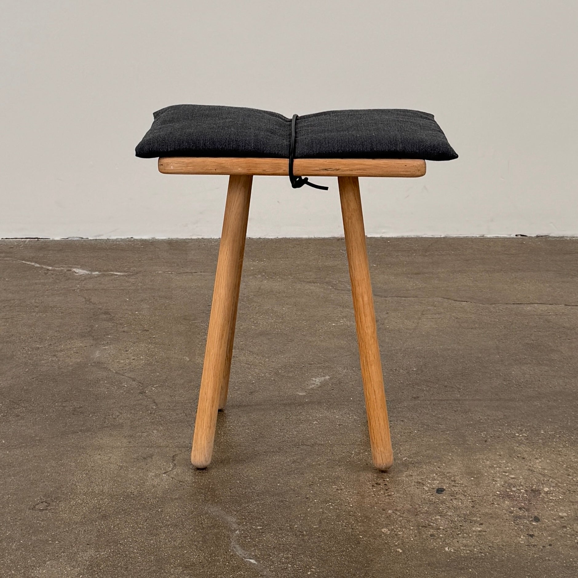 A Fritz Hansen Skagerak Georg Stool, featuring three wooden legs and a padded black cushion on top, is photographed against a plain white background.