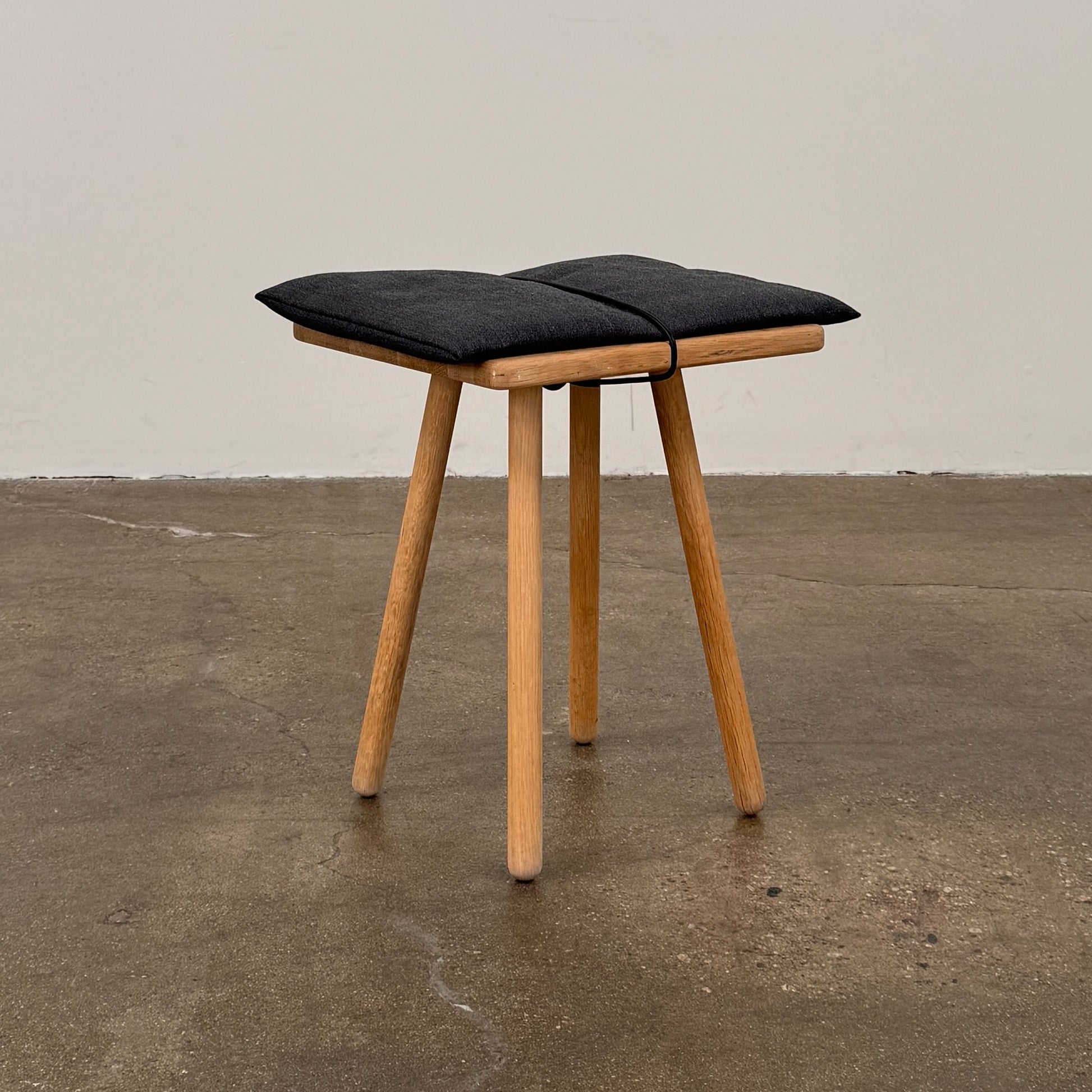 A Fritz Hansen Skagerak Georg Stool, featuring three wooden legs and a padded black cushion on top, is photographed against a plain white background.
