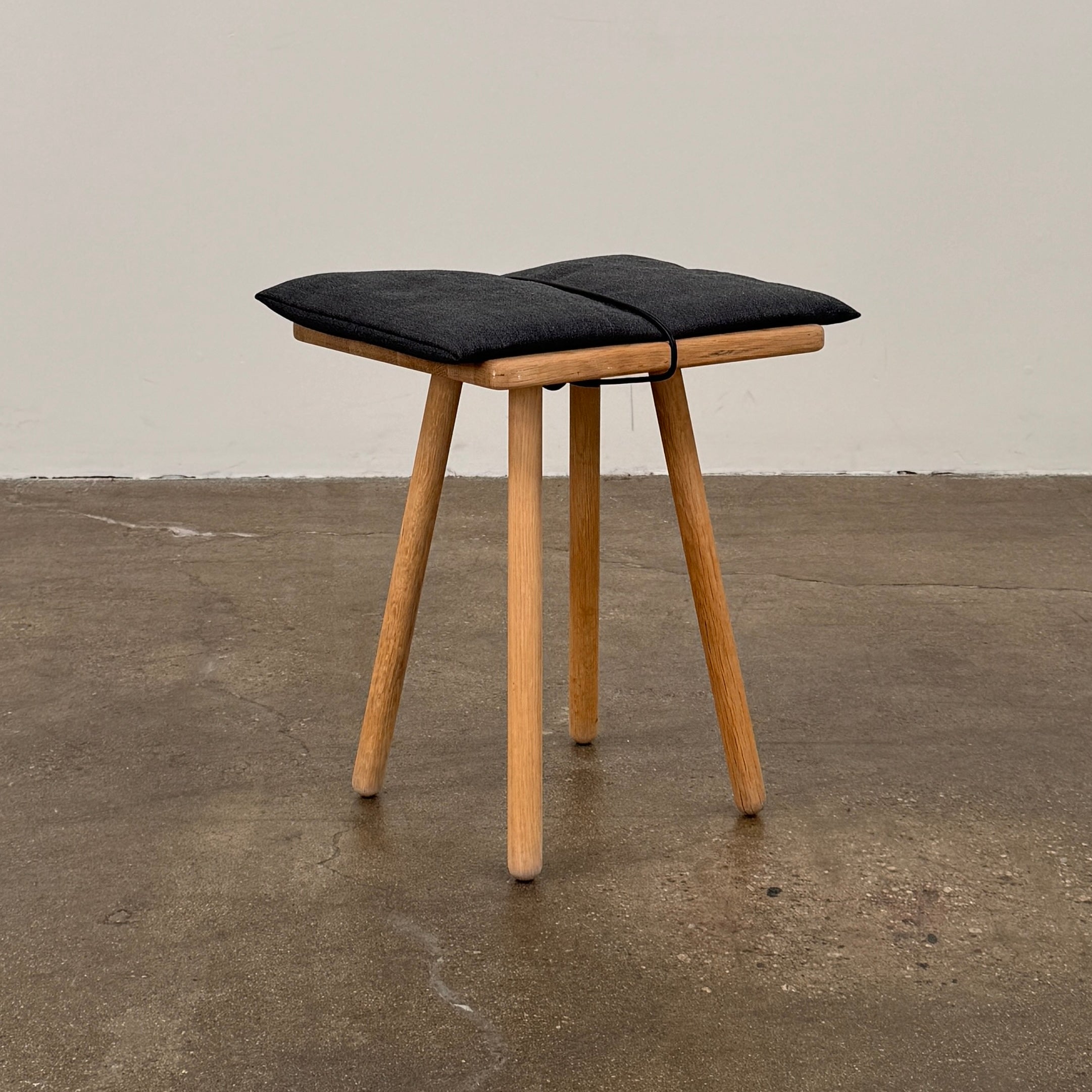 A Fritz Hansen Skagerak Georg Stool, featuring three wooden legs and a padded black cushion on top, is photographed against a plain white background.