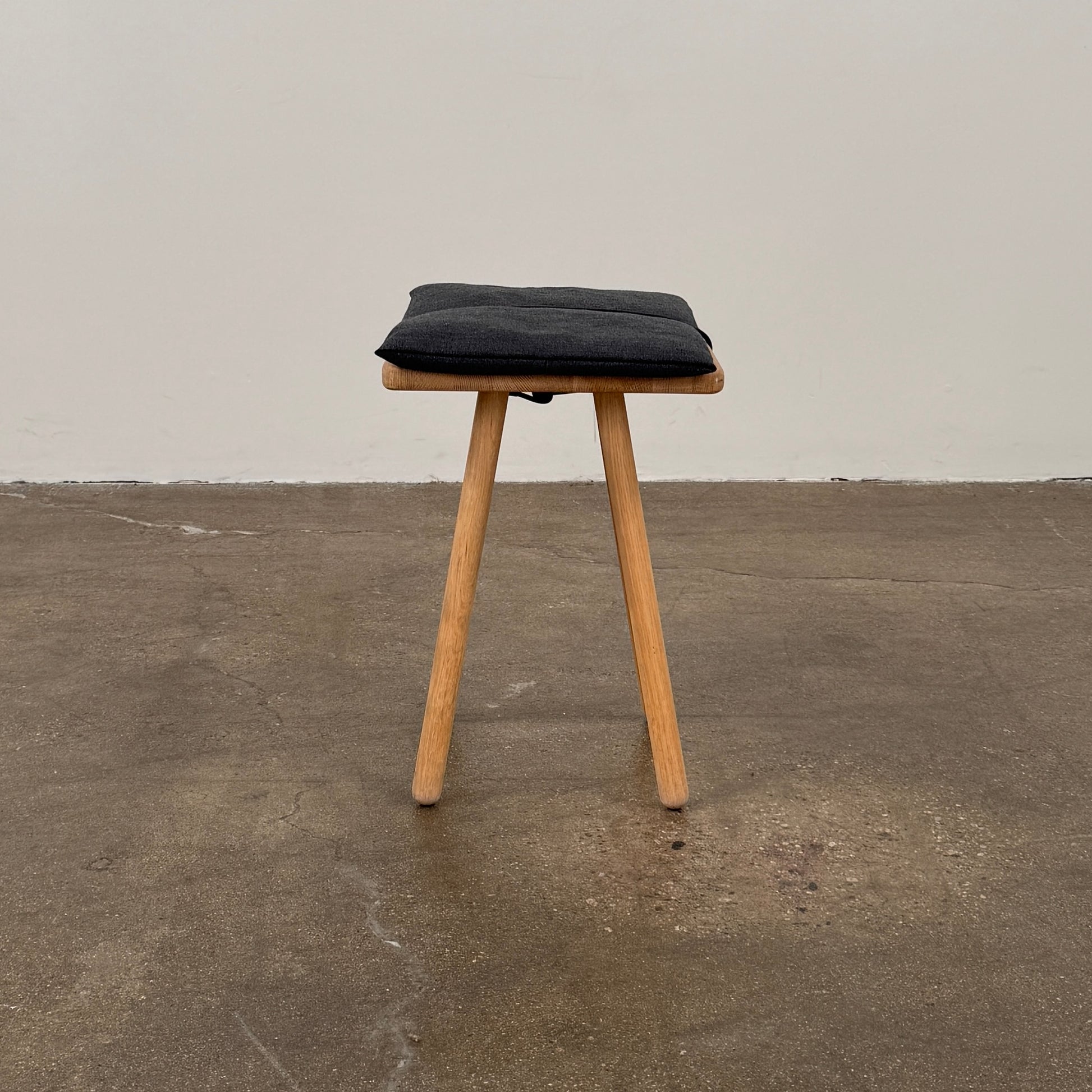 A Fritz Hansen Skagerak Georg Stool, featuring three wooden legs and a padded black cushion on top, is photographed against a plain white background.