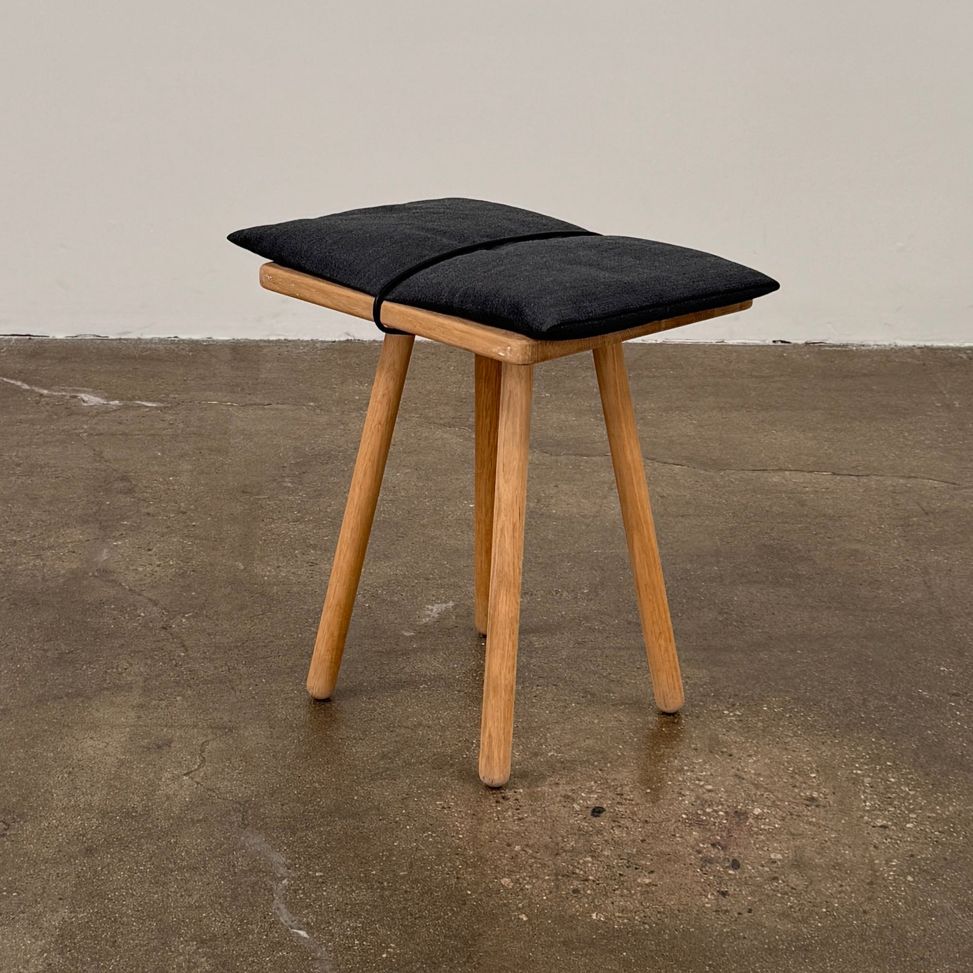 A Fritz Hansen Skagerak Georg Stool, featuring three wooden legs and a padded black cushion on top, is photographed against a plain white background.
