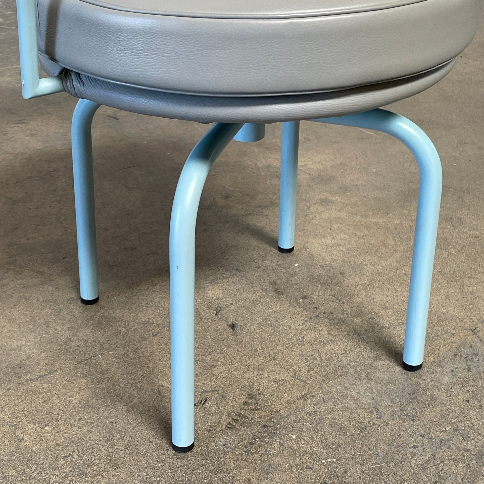 A gray leather Cassina LC7 Swivel Chair, featuring a circular seat, tubular backrest, and light blue metal legs, embodies modern design by Le Corbusier on a white background.