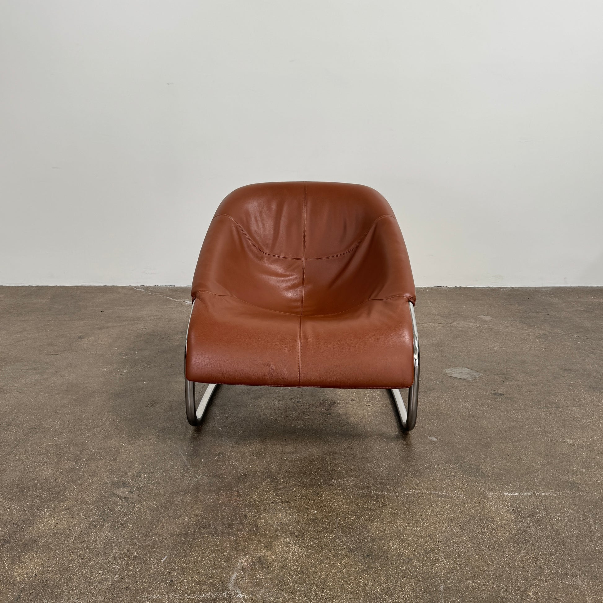 The Minotti Cortina Armchair by Minotti features luxurious brown leather upholstery and a gracefully curved seat, supported by a sleek chrome metal frame, and is showcased against a plain white background.