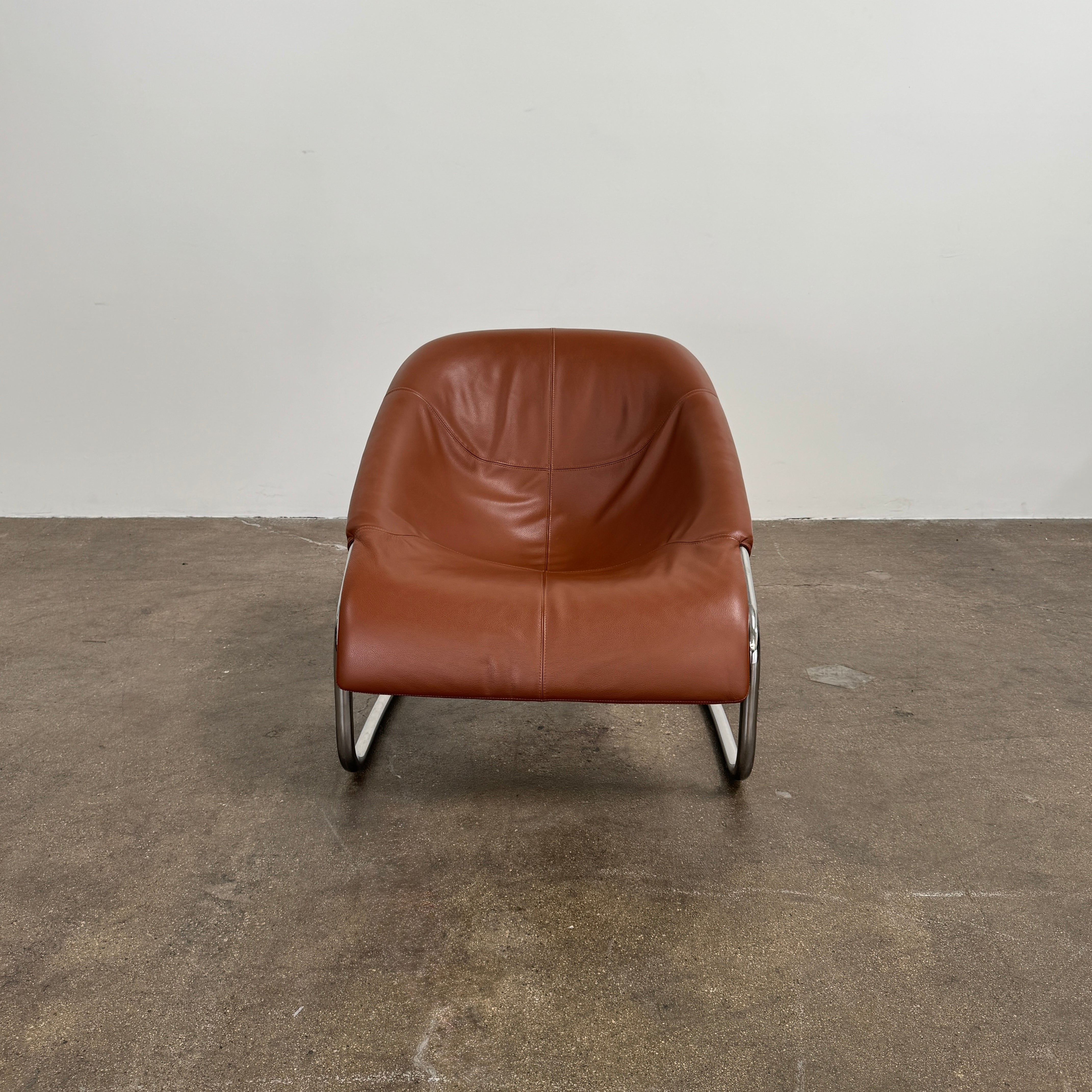 The Minotti Cortina Armchair by Minotti features luxurious brown leather upholstery and a gracefully curved seat, supported by a sleek chrome metal frame, and is showcased against a plain white background.