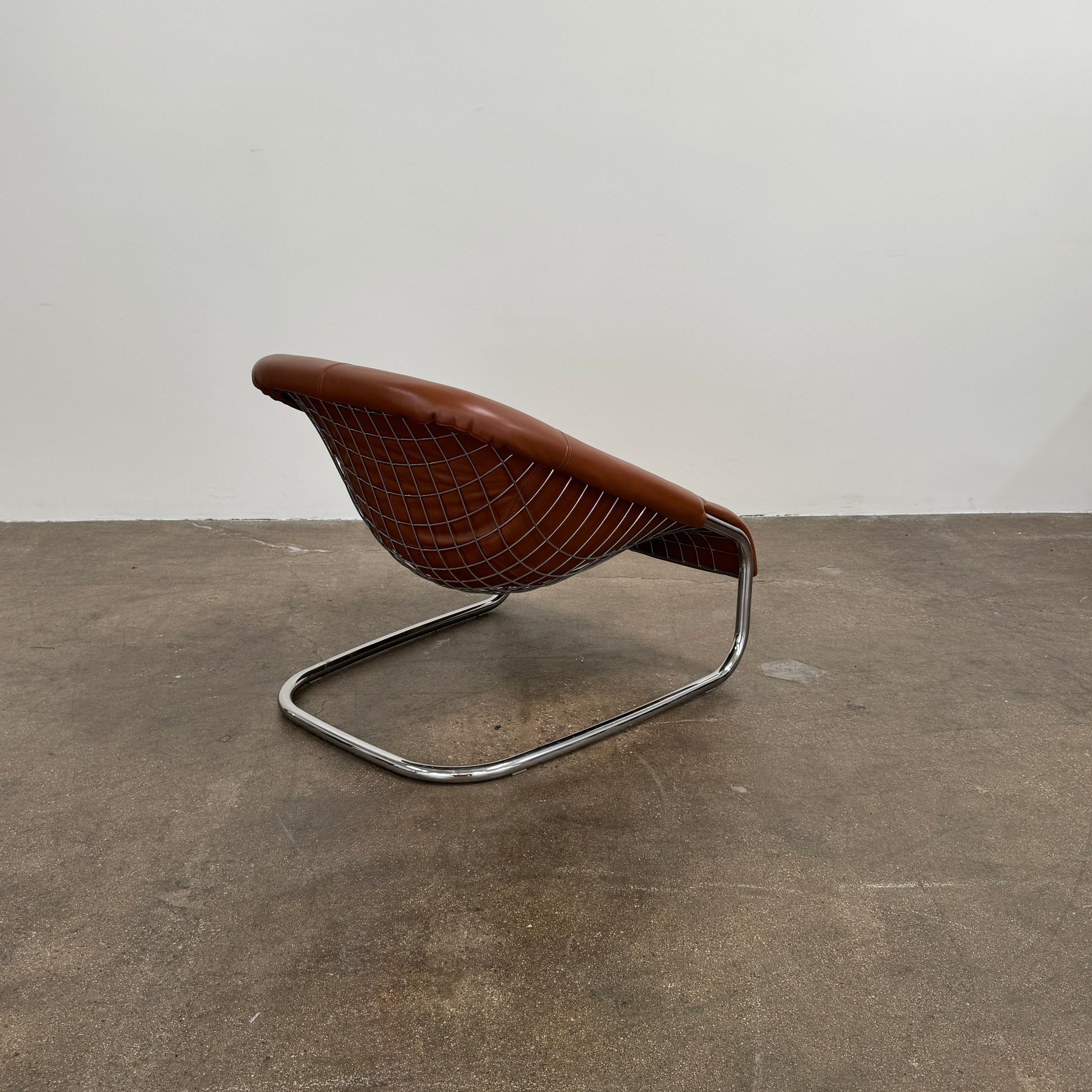 The Minotti Cortina Armchair by Minotti features luxurious brown leather upholstery and a gracefully curved seat, supported by a sleek chrome metal frame, and is showcased against a plain white background.