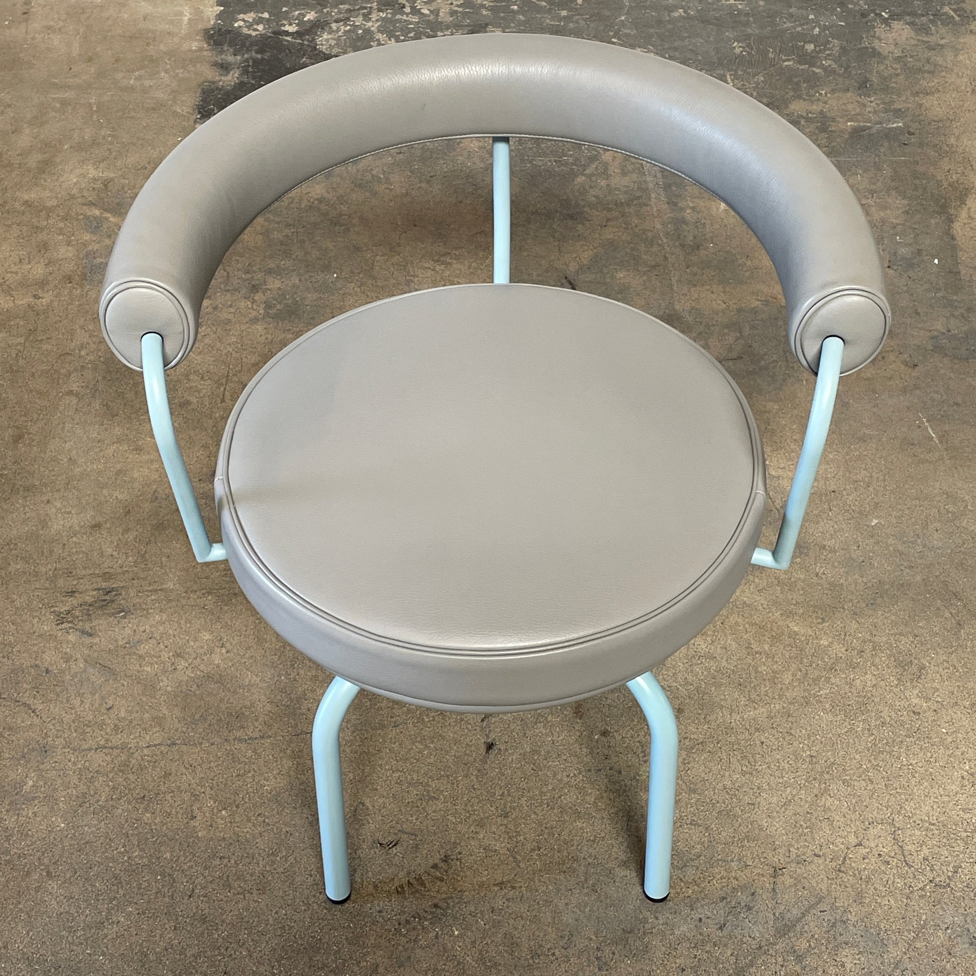 A gray leather Cassina LC7 Swivel Chair, featuring a circular seat, tubular backrest, and light blue metal legs, embodies modern design by Le Corbusier on a white background.