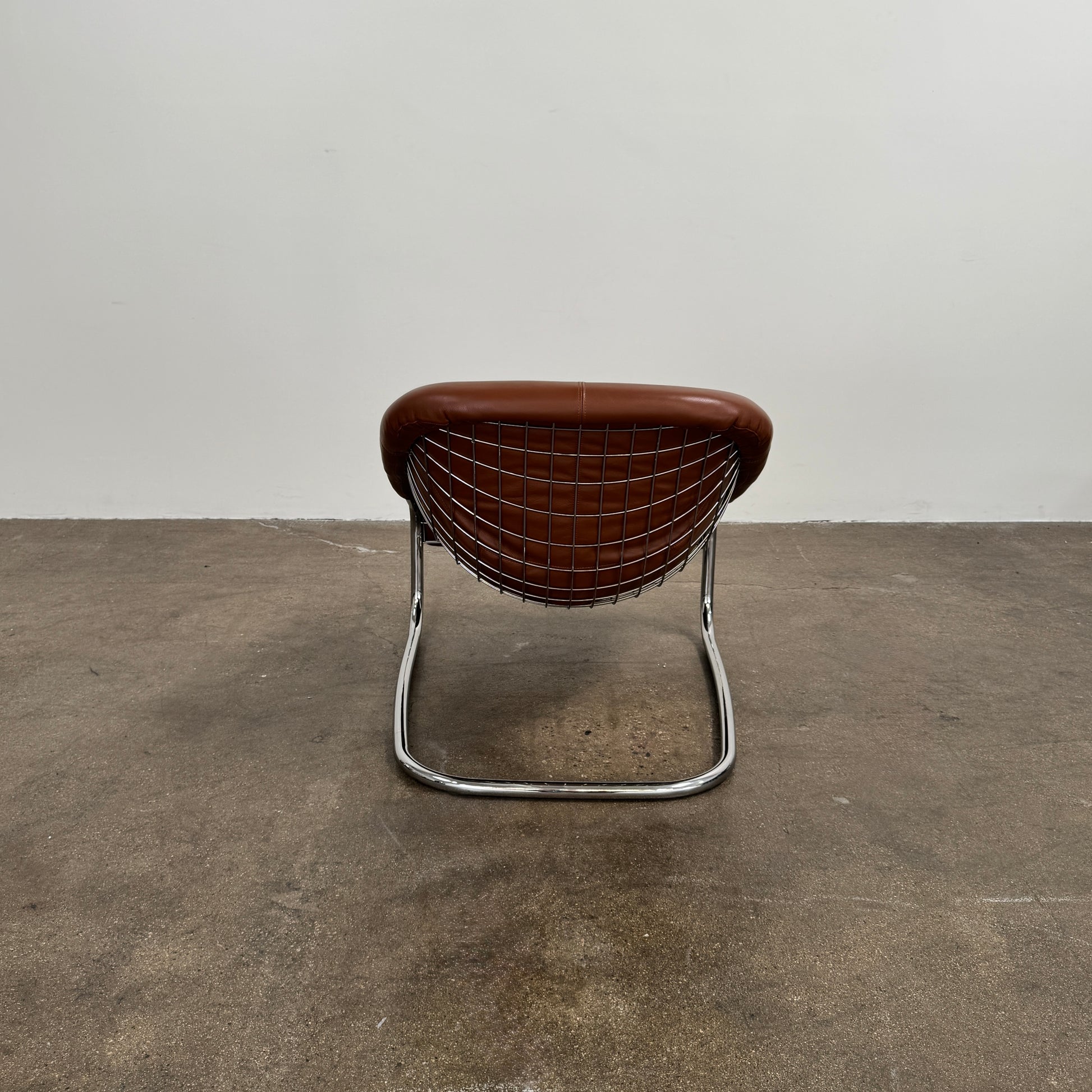 The Minotti Cortina Armchair by Minotti features luxurious brown leather upholstery and a gracefully curved seat, supported by a sleek chrome metal frame, and is showcased against a plain white background.