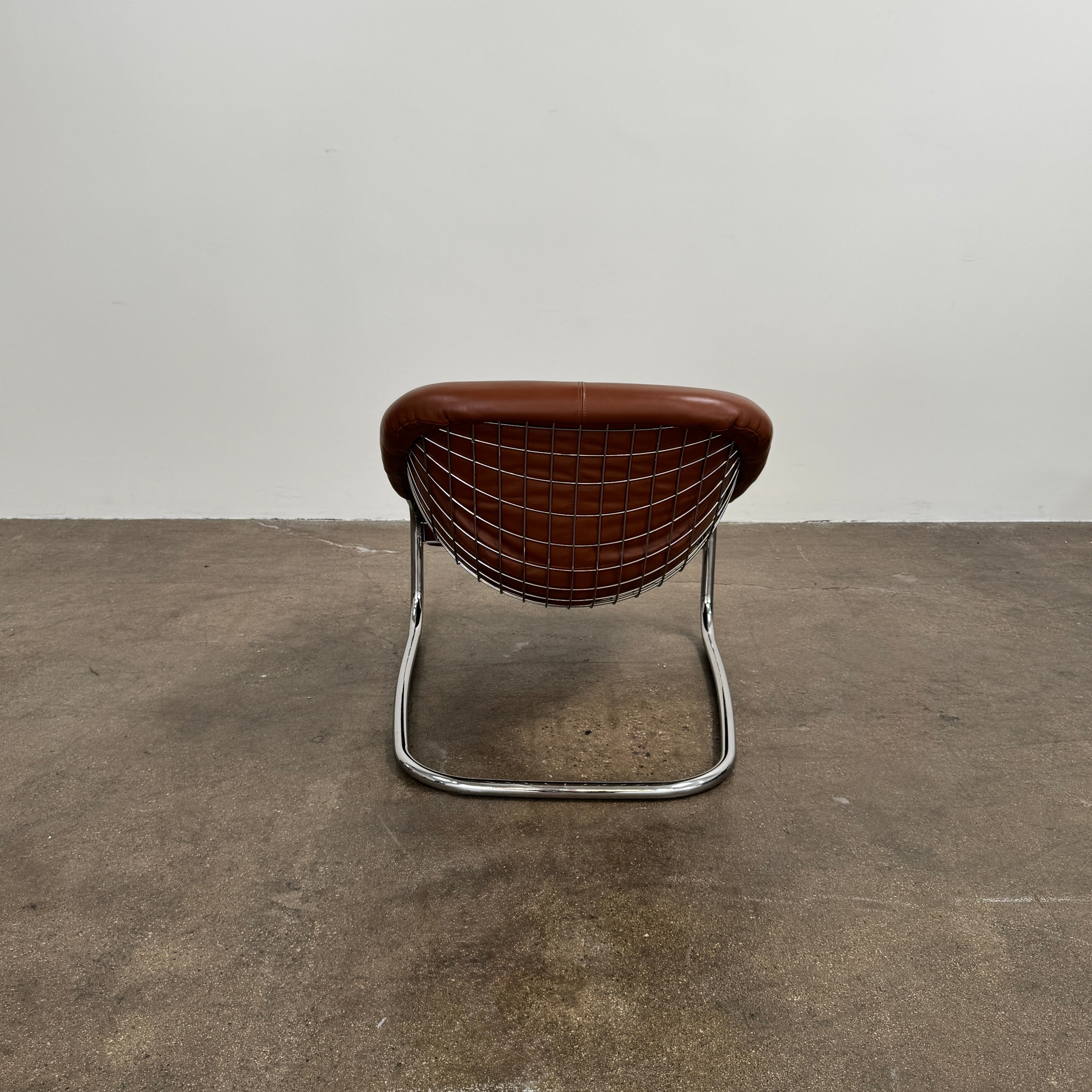 The Minotti Cortina Armchair by Minotti features luxurious brown leather upholstery and a gracefully curved seat, supported by a sleek chrome metal frame, and is showcased against a plain white background.