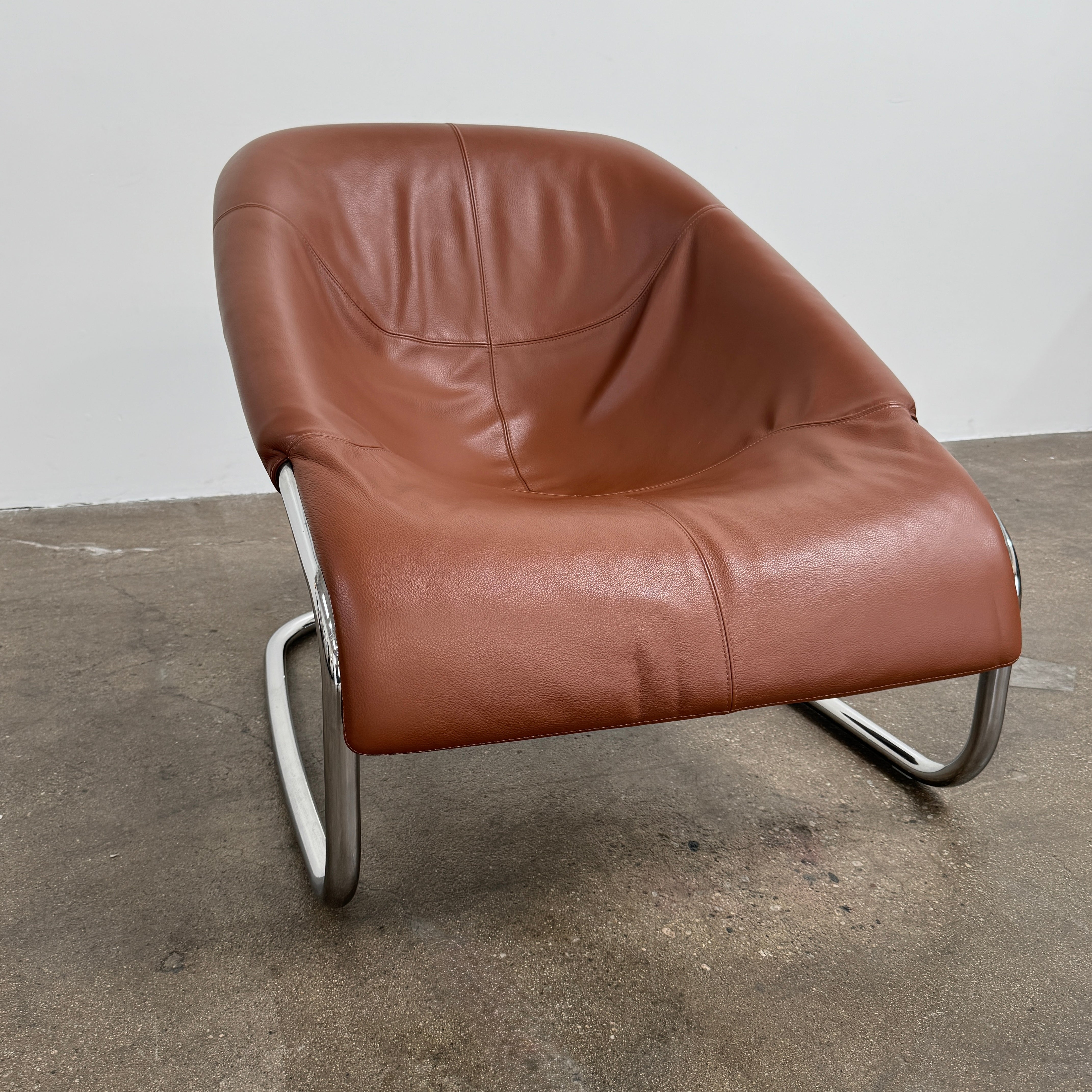 The Minotti Cortina Armchair by Minotti features luxurious brown leather upholstery and a gracefully curved seat, supported by a sleek chrome metal frame, and is showcased against a plain white background.