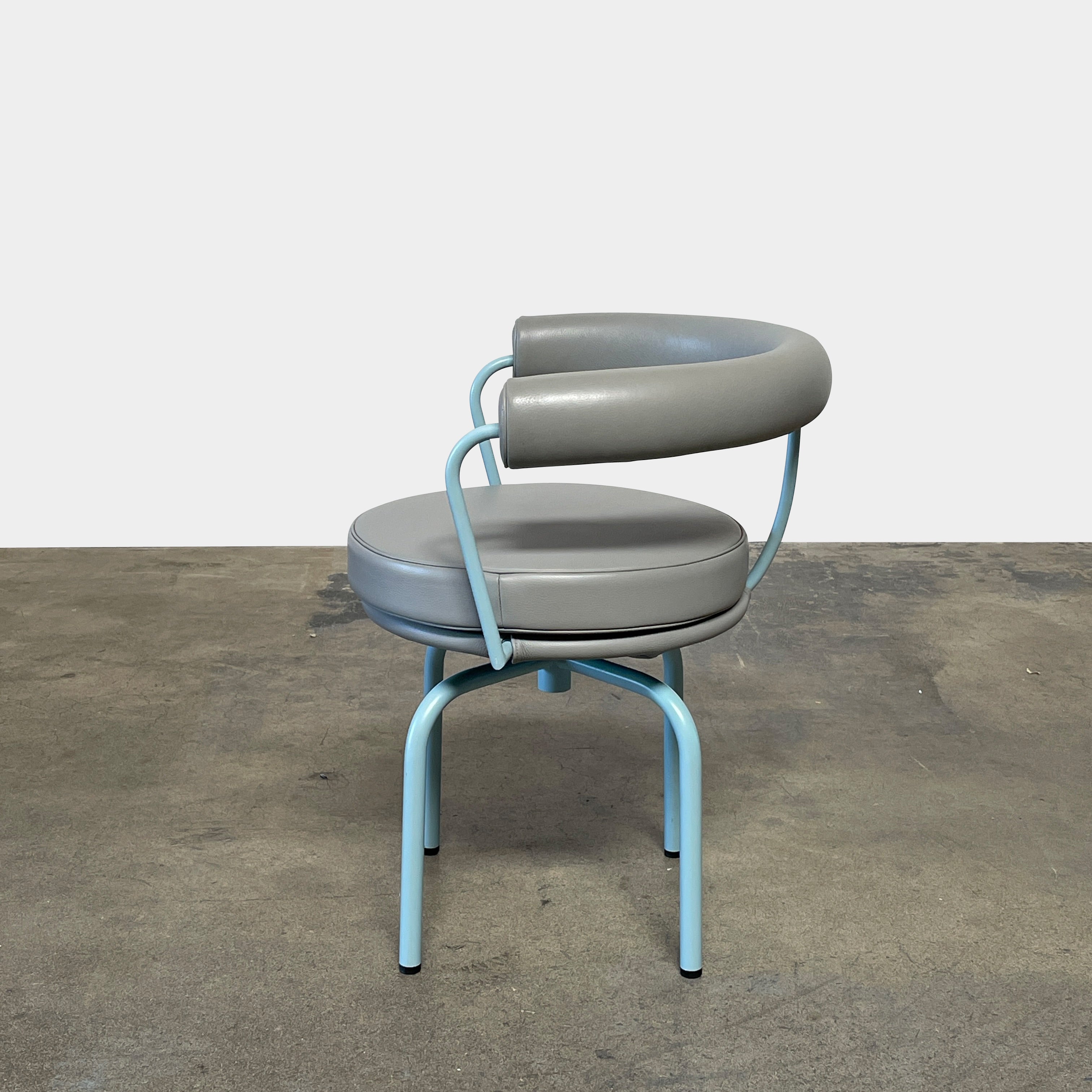 A gray leather Cassina LC7 Swivel Chair, featuring a circular seat, tubular backrest, and light blue metal legs, embodies modern design by Le Corbusier on a white background.
